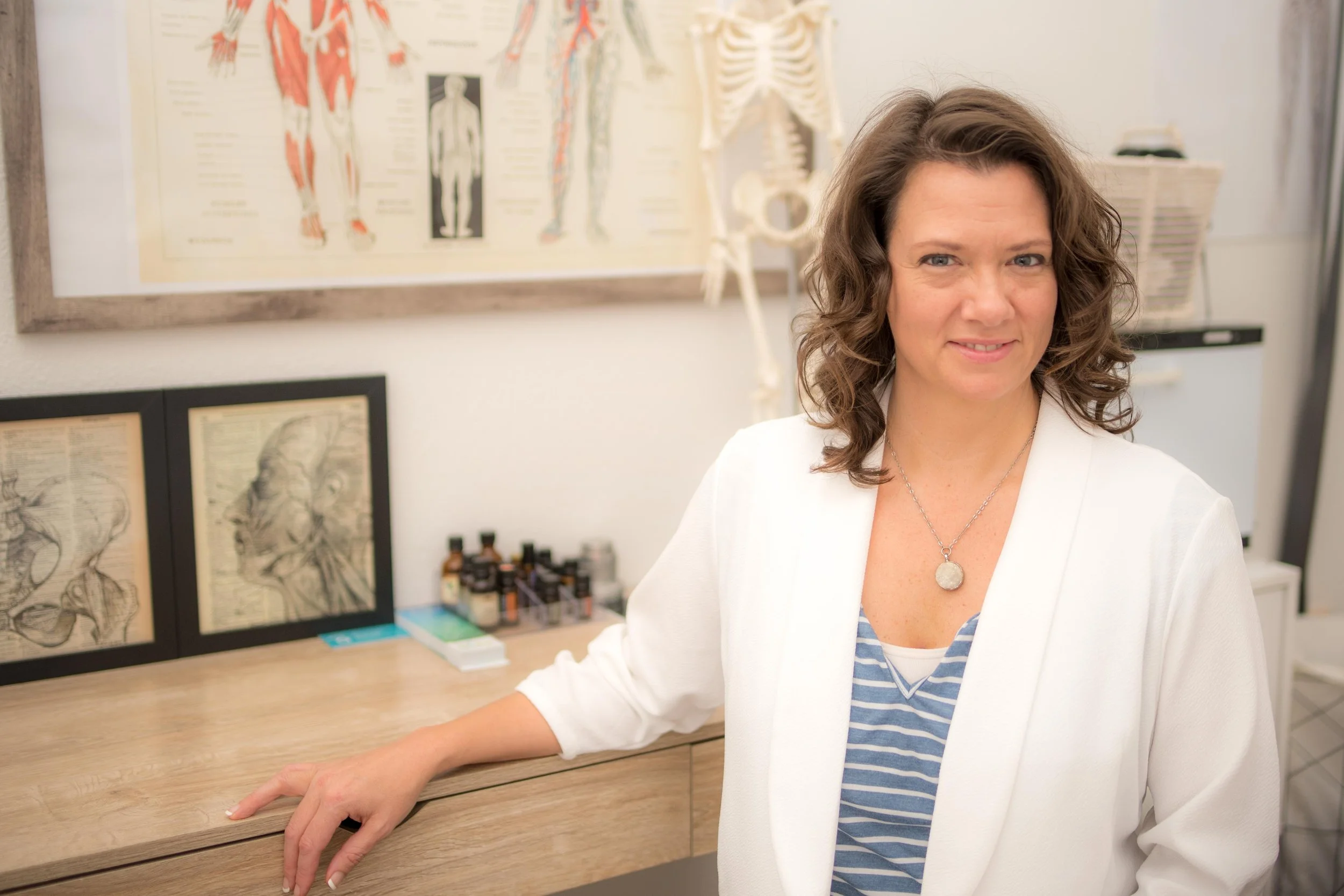 A woman with curly brown hair sitting at a wooden desk in a medical office. Behind her are anatomical charts and medical illustrations including a human skeleton and muscle diagrams.