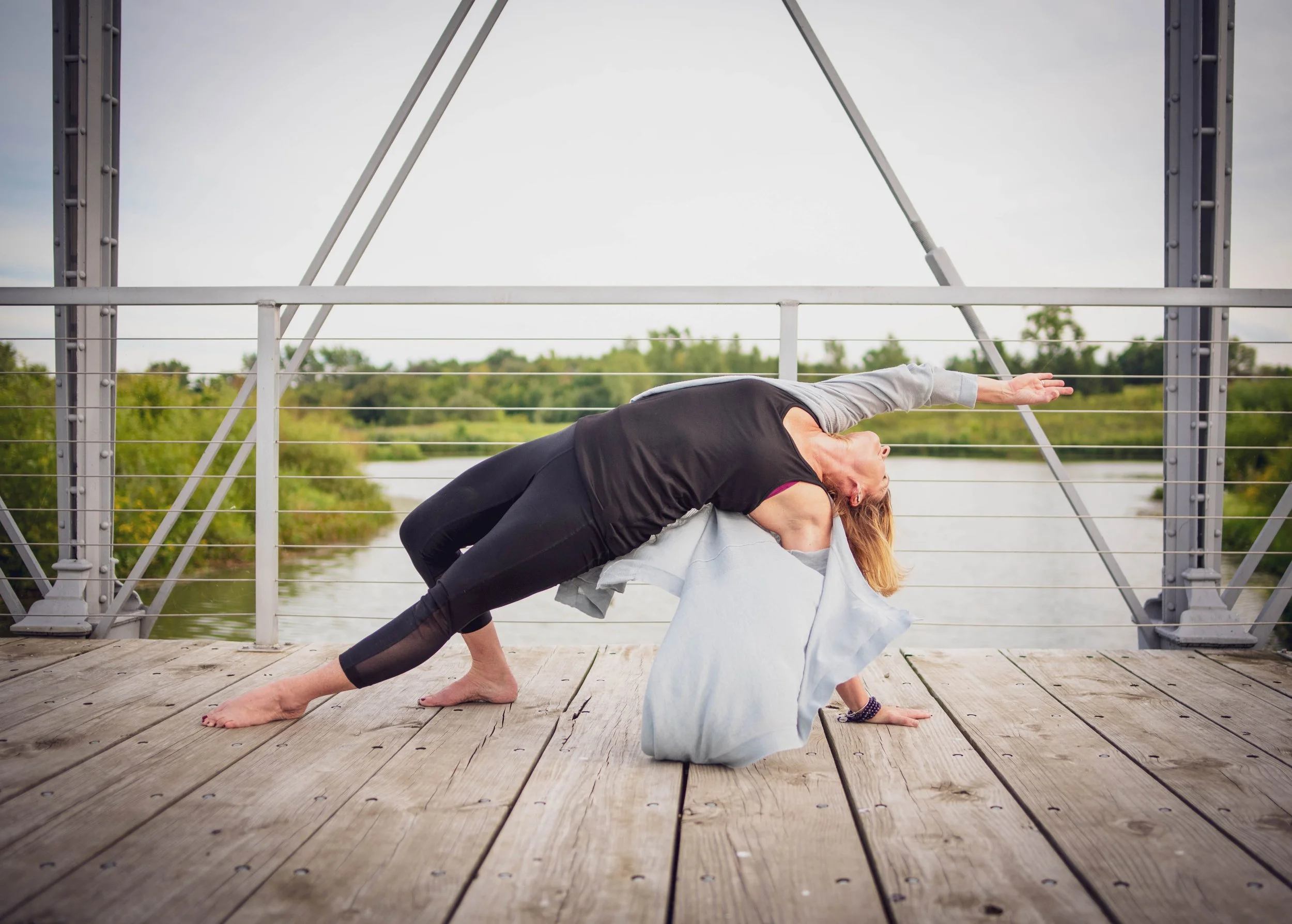 A person practicing yoga on a bridge over a river, performing a side stretch pose with one arm extending over the head and the other supporting on the wooden bridge surface.