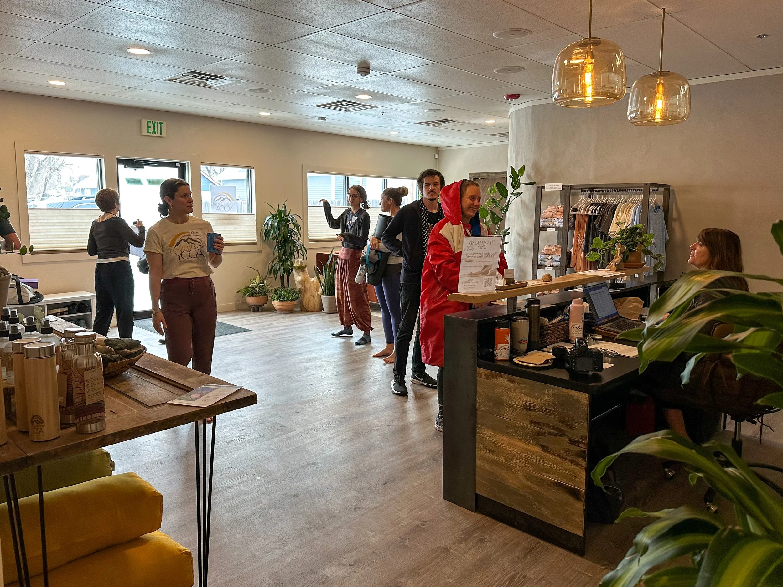 People standing in line at a reception desk in a well-lit yoga studio, with plants and clothing on display in the background.