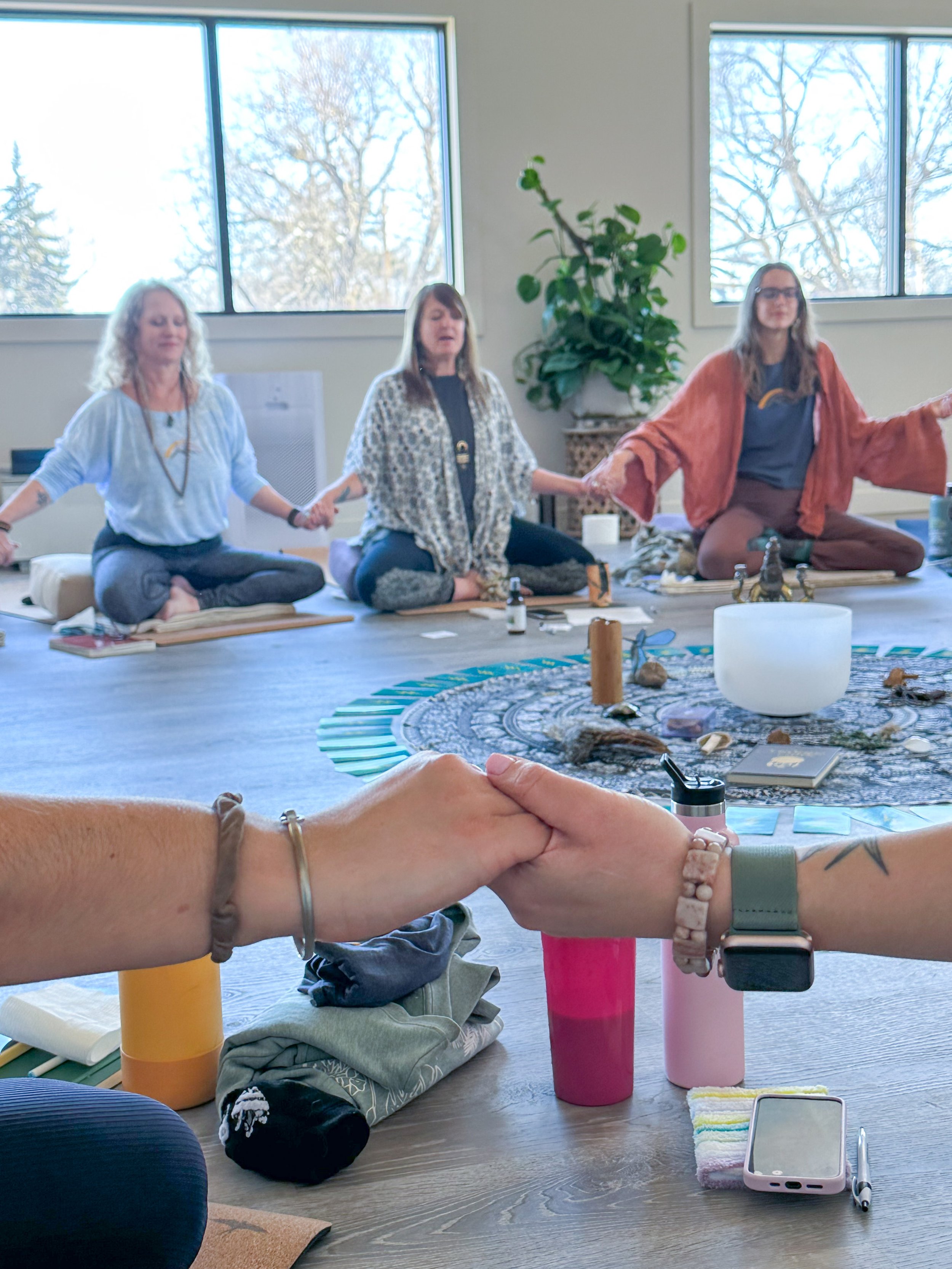 A group of women sitting cross-legged in meditation pose on mats, holding hands, in a bright room with large windows and a decorative circle of objects and candles in the center.