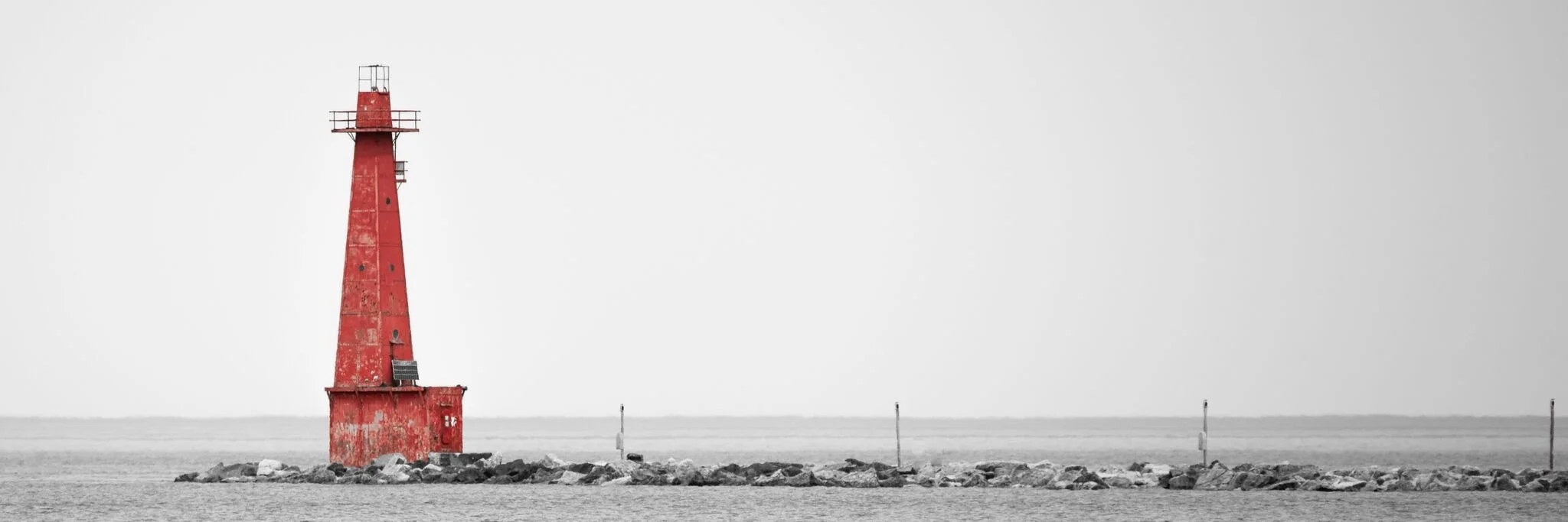 Red lighthouse on a rocky breakwater against a cloudy sky, black and white surroundings.