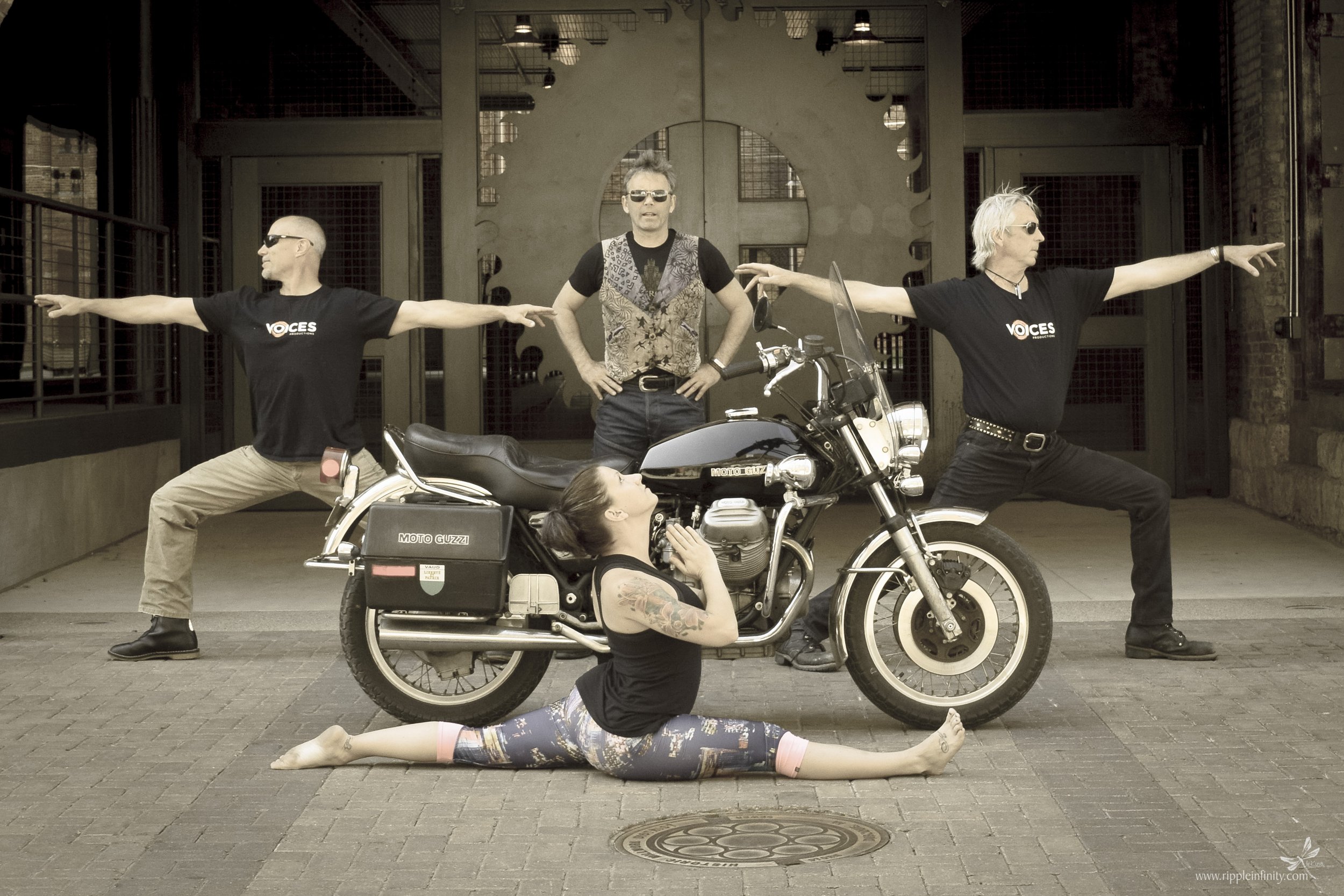 Group of five people, three men and two women, posing with a motorcycle in an urban setting. One woman is performing a split on the ground in front of the motorcycle, while two men extend their arms outward in yoga-like poses on either side of the mo