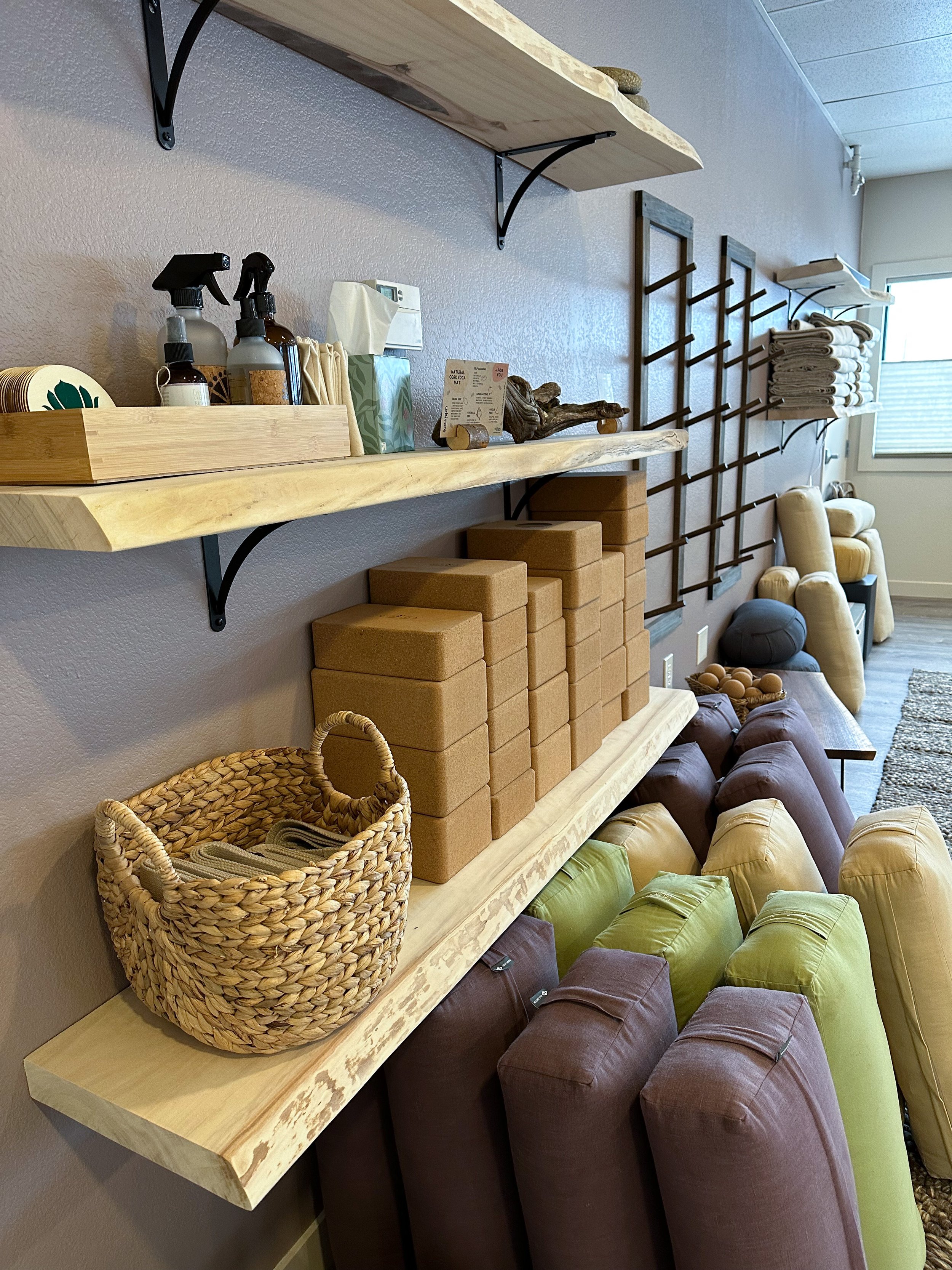 View of a cozy room with wooden shelves mounted on a beige wall, holding various items including cleaning spray bottles, tissue box, cakes, and decor. Below the shelves are stacked brown bricks, a woven basket, and colorful cushions and pillows on a 