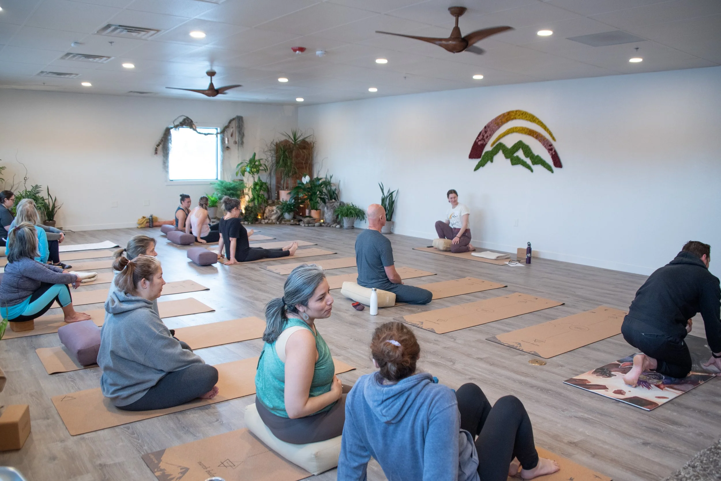 People participating in a yoga class or meditation session in a spacious, well-lit room with wooden flooring, potted plants in the background, and a colorful mountain logo on the wall.