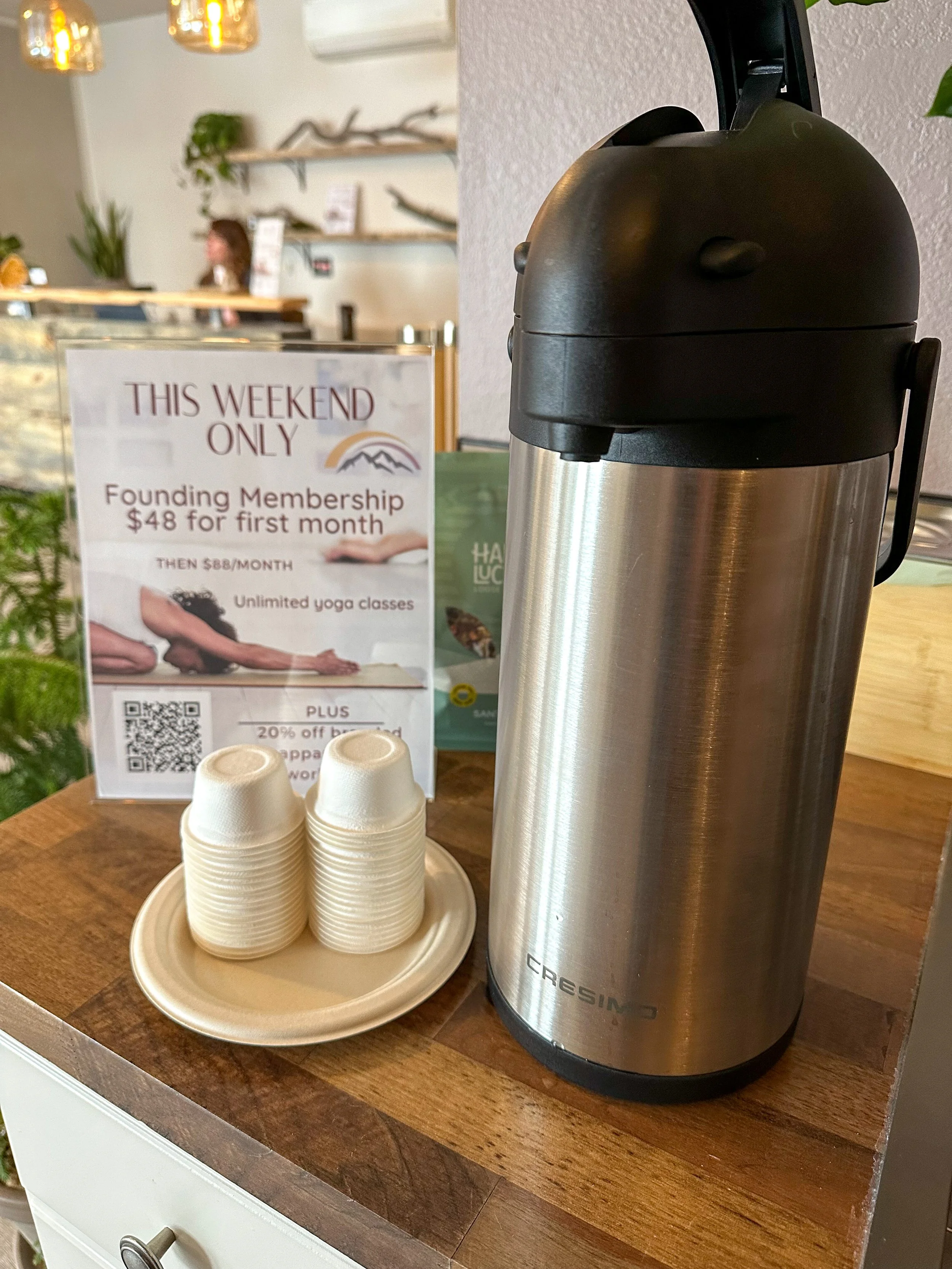 A stainless steel coffee carafe on a wooden table next to a sign promoting a new membership offer at a yoga studio, with disposable cups on a small plate and a background of a reception area.
