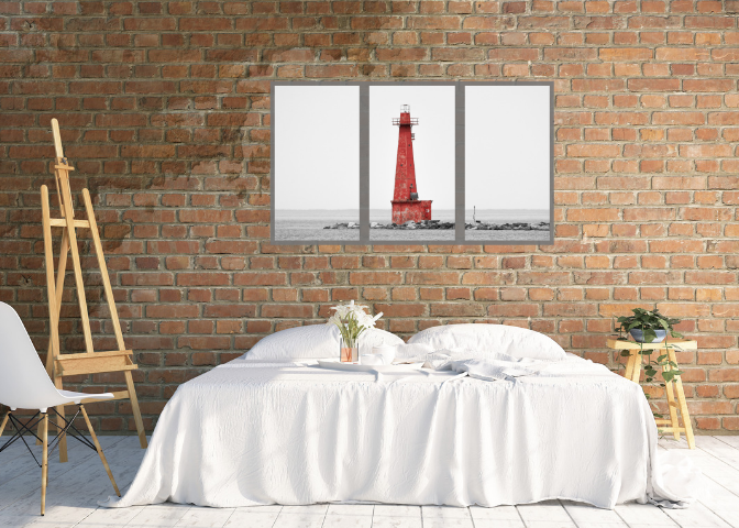 A bedroom with a brick wall, a white bed with white bedding, a small table with a plant, a chair, and a three-panel picture of a red lighthouse on the wall.