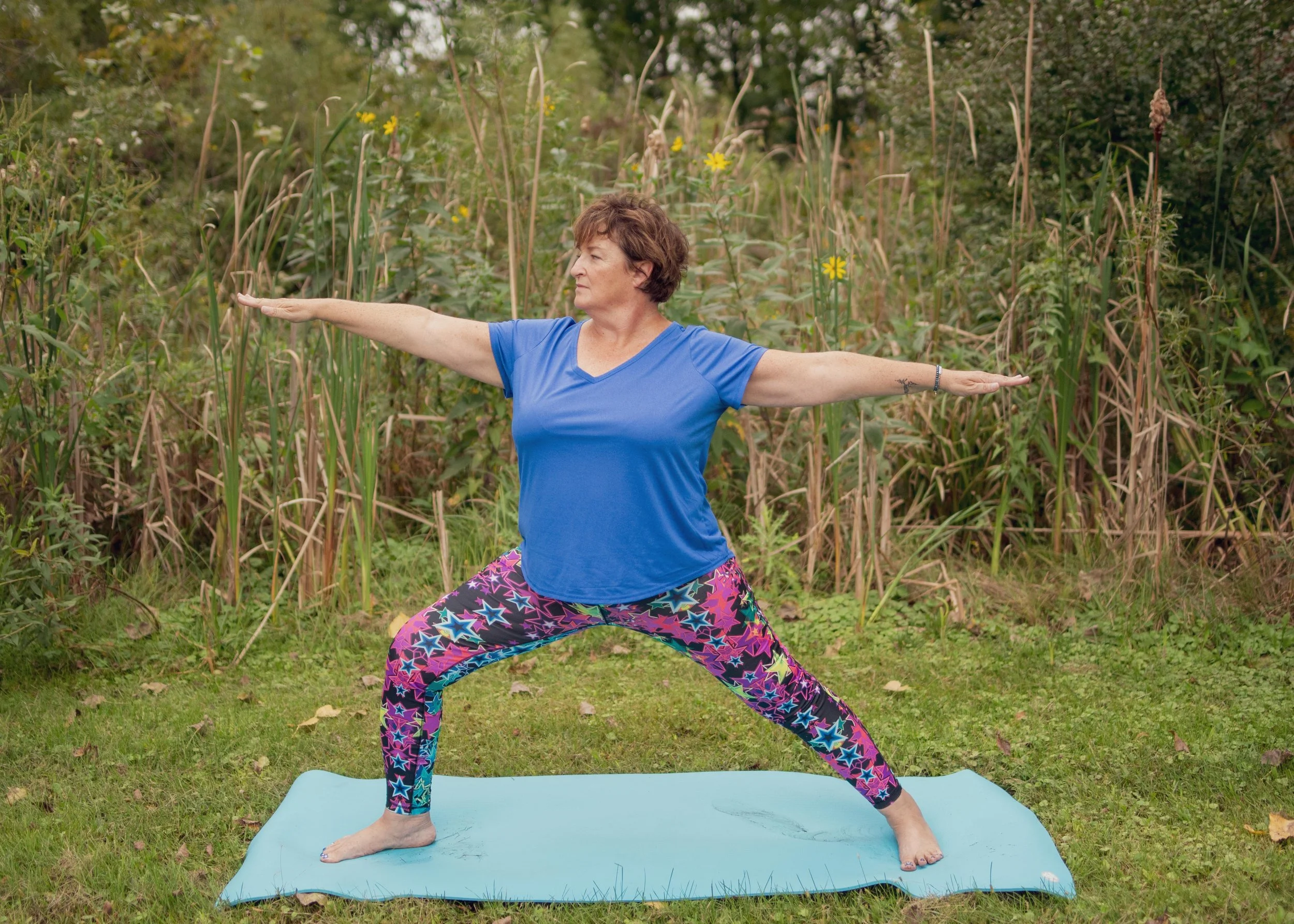 A woman practicing yoga outdoors on a blue yoga mat, wearing a blue t-shirt and colorful star-patterned leggings, standing on grass with her arms extended horizontally, in front of tall grass and yellow flowers.