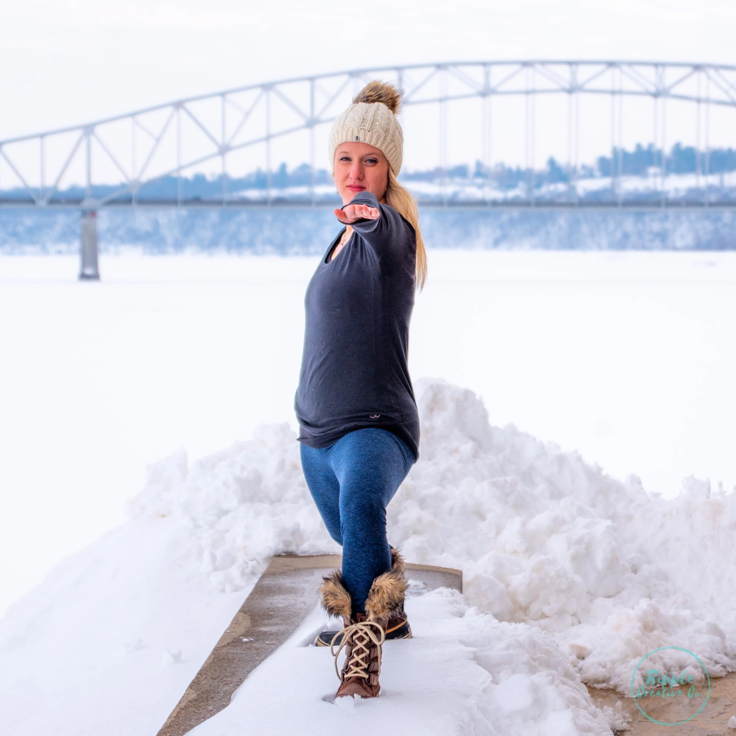 Woman in winter clothes performing a yoga pose outdoors in the snow with a bridge in the background.