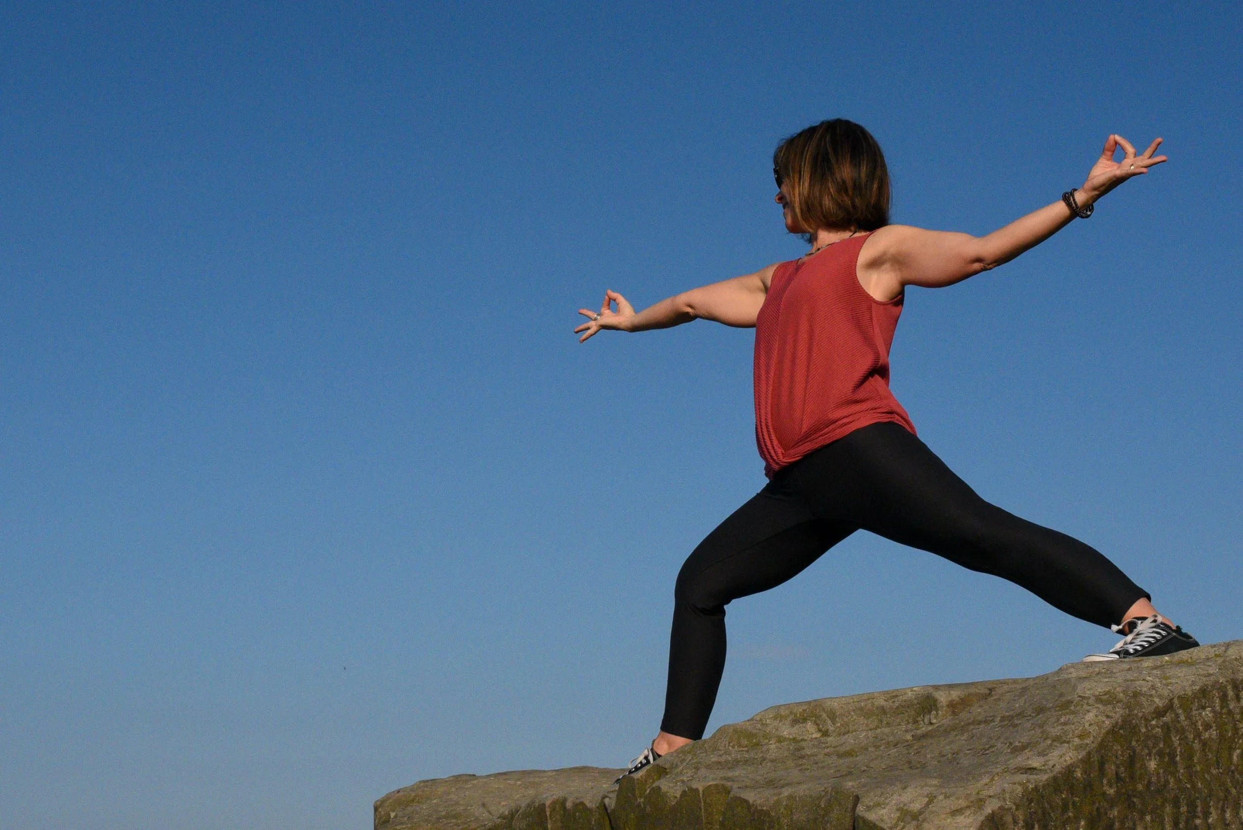 A woman practicing yoga outdoors on a rocky surface against a clear blue sky, standing with arms extended and one leg bent in a Warrior II pose.