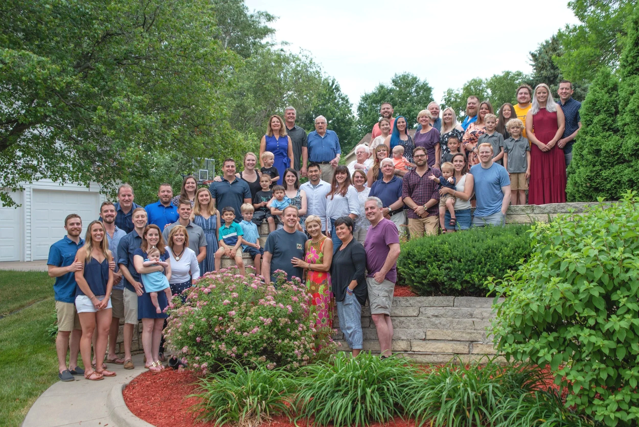 A large group of people gathered outdoors on a staircase in a garden, smiling for a group photo, surrounded by trees and bushes.