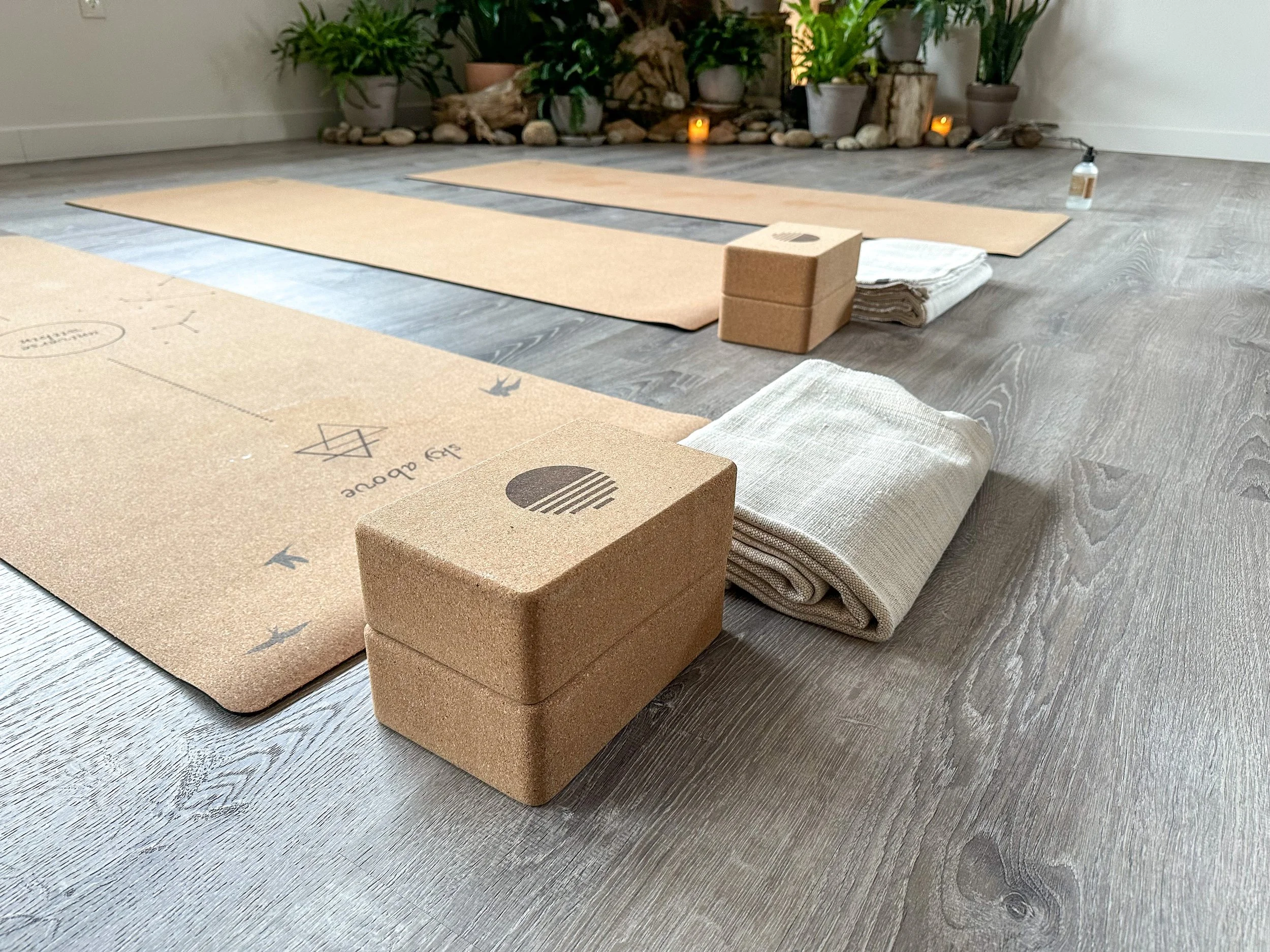 Yoga mats, blocks, and props arranged on a wooden floor in a serene indoor space with plants and candles in the background.