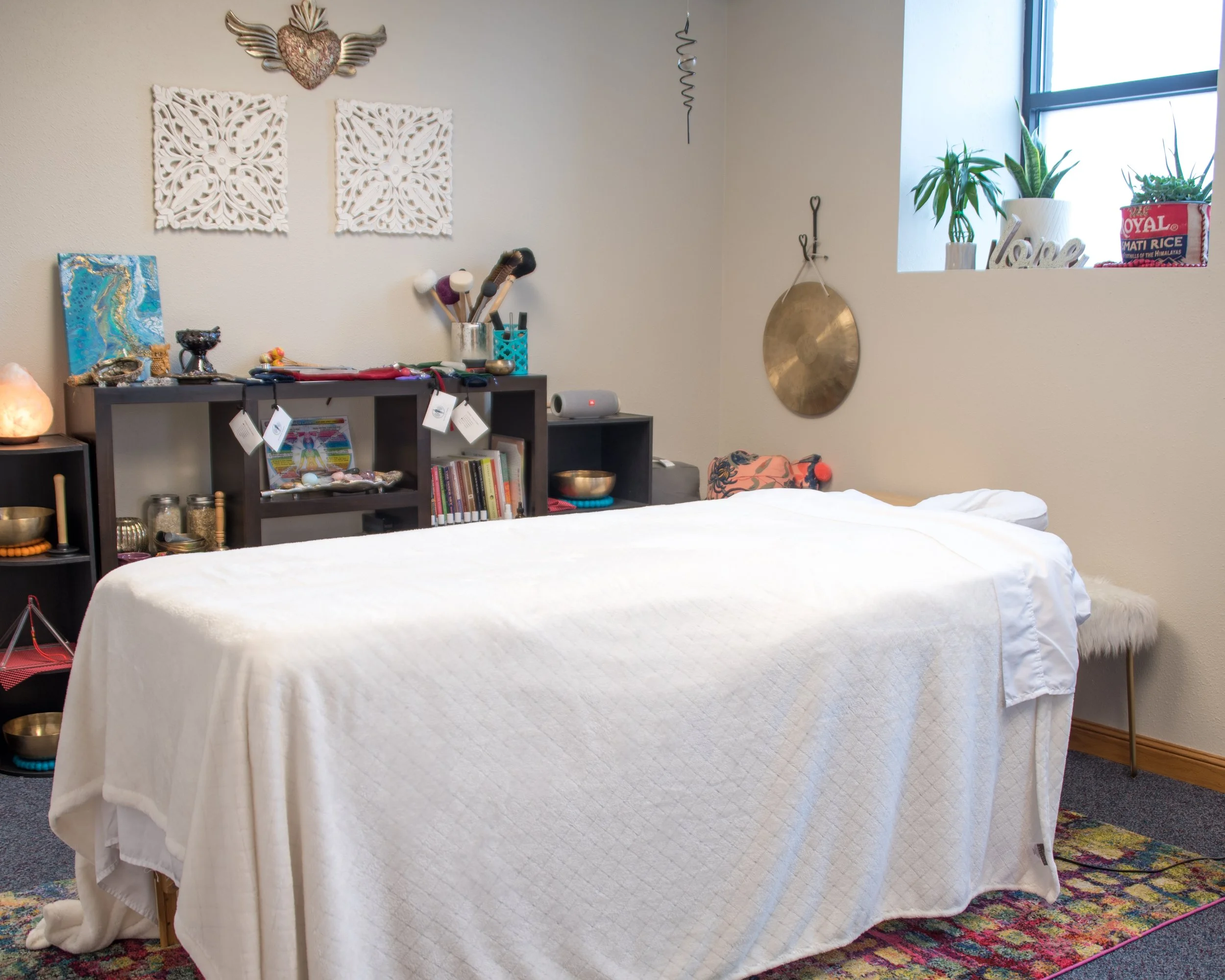 A massage therapy room with a massage table covered in a white sheet. Behind the table are shelves with various decorative items, books, and bowls. The wall has wall art and a large gong, with a window above the shelf holding potted plants and decora