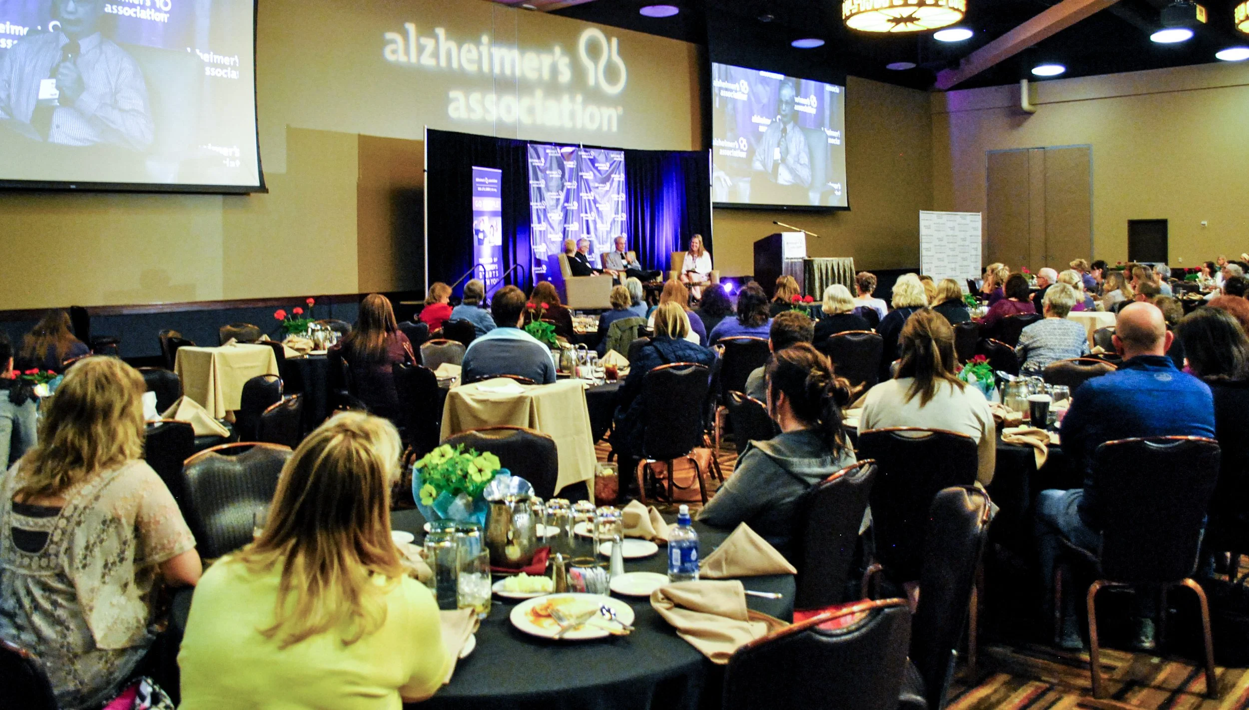 A conference room filled with seated attendees listening to a panel discussion on stage, with large screens displaying the 'Alzheimer's Association' logo.