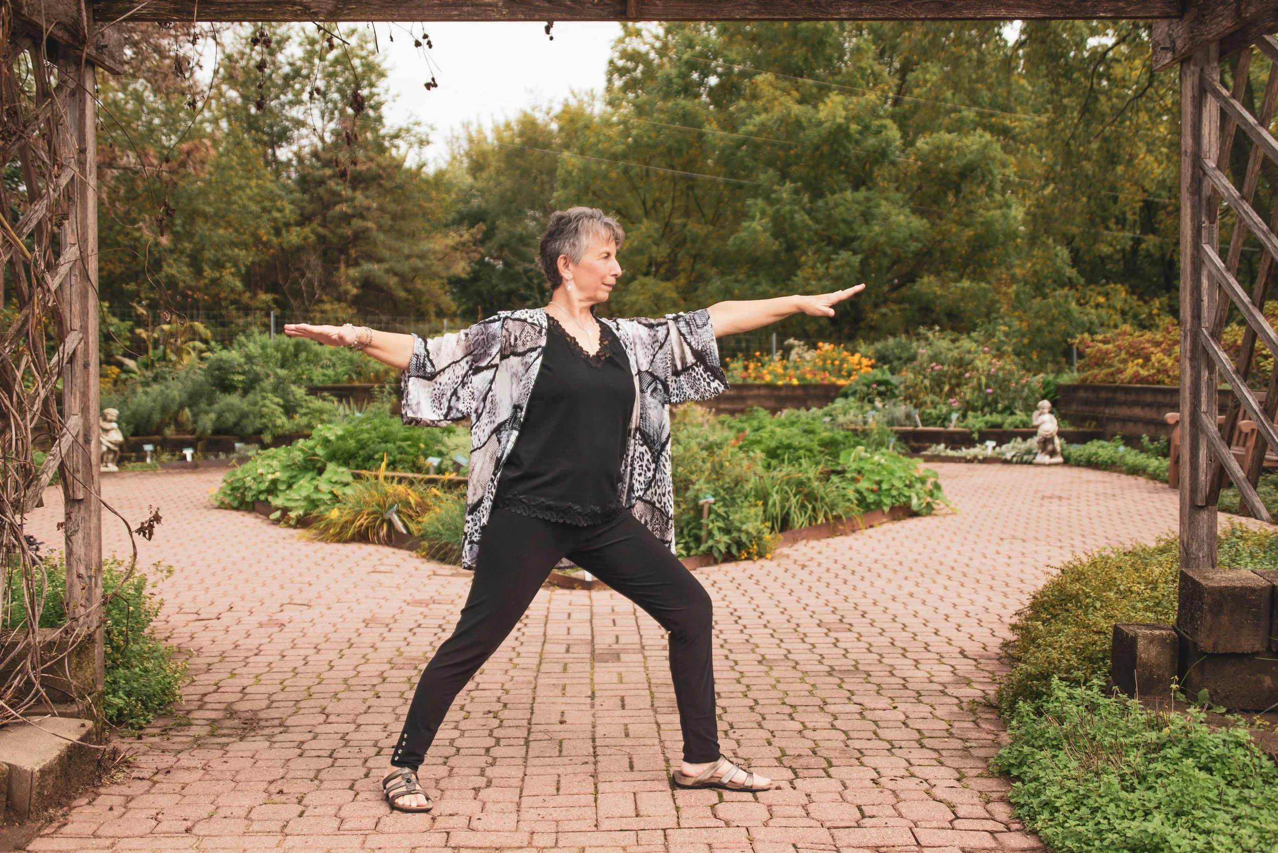 A woman practicing yoga outdoors on a brick path in a garden with trees and flowers.
