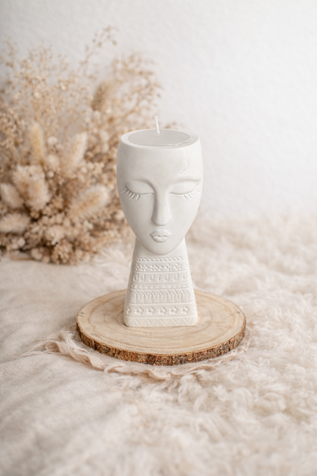 Weiße Kerze in Form eines Gesichtskopfes auf einem Holzstück mit dekorierender Muster, im Hintergrund beige Trockenblumen, auf einer weichen, cremefarbenen Decke
