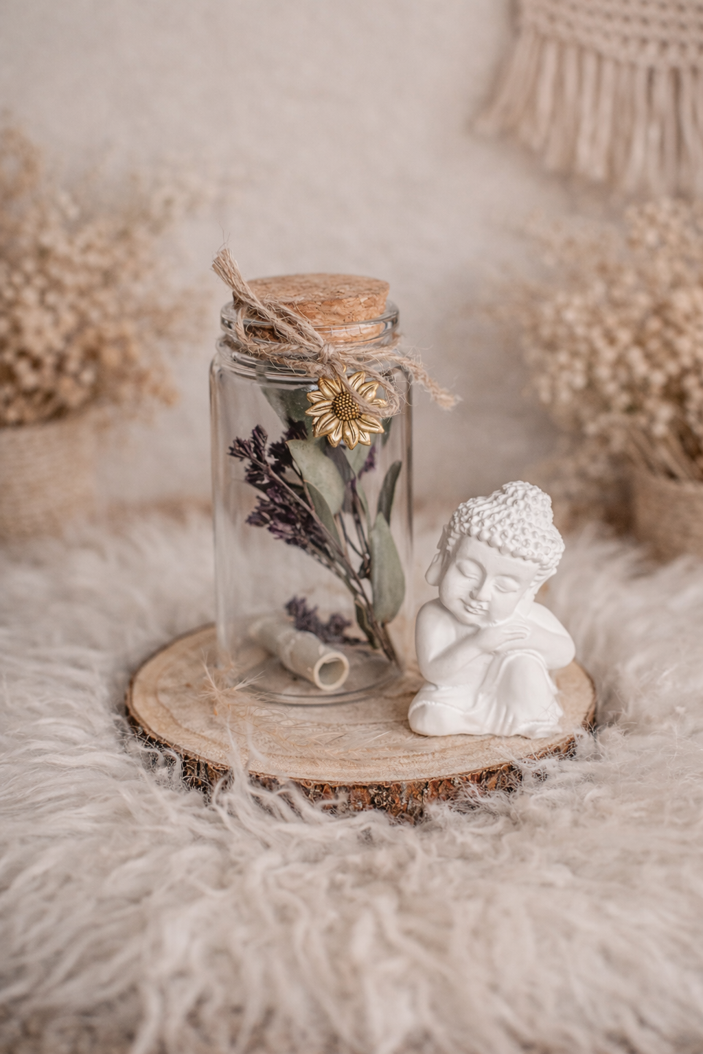 Eine kleine Buddha-Figur aus weißem Material, sitzend mit verschränkten Armen auf einer runden Holzscheibe, neben einer Glaskaraffe mit getrockneten Blumen und einer goldfarbenen Sonnenblumen-Decor. Im Hintergrund beige und braune Bast- und Filzdekorationen.