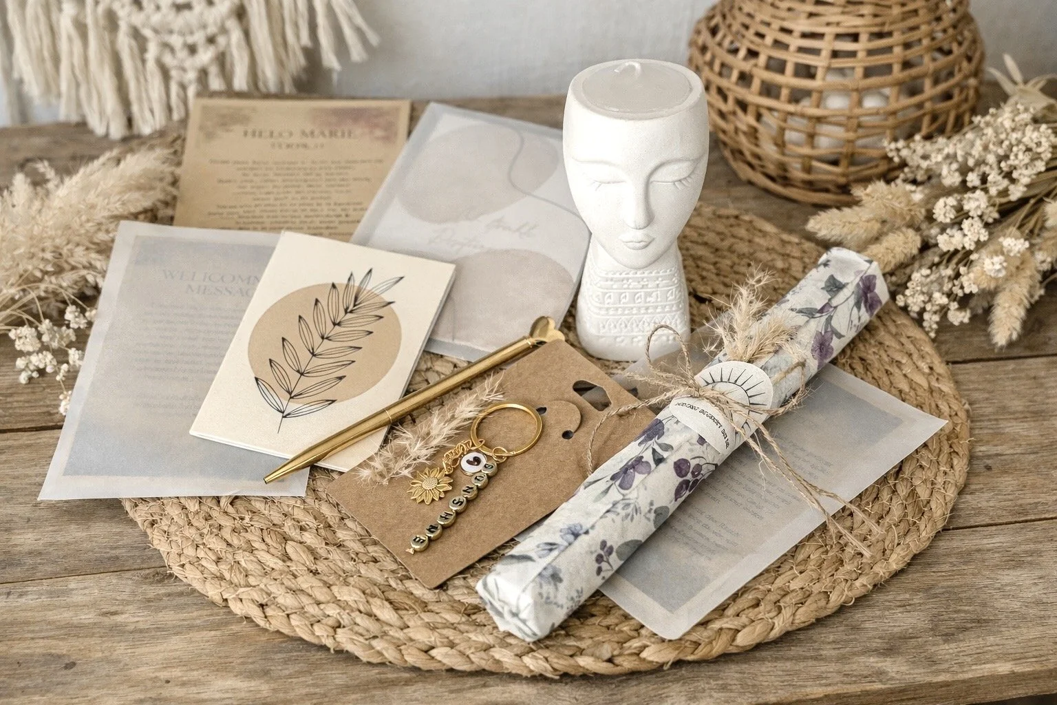 Verschiedene Geschenke und Dekorationen auf einem runden, geflochtenen Untersetzer, darunter eine weiße Porzellanfigur, Briefumschläge, eine goldene Stift, eine Schlüsselanhänger, ein Geschenk mit Lavendelmuster und getrockneten Blumen.