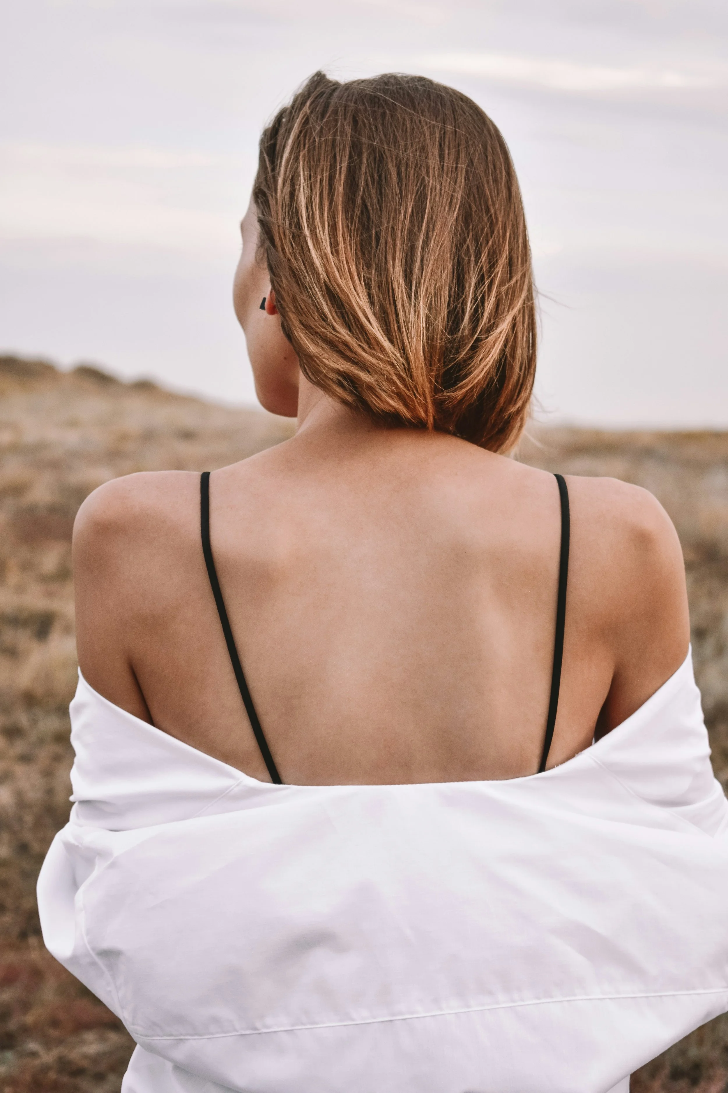 Back view of a woman with shoulder-length brown hair, wearing a white off-shoulder shirt and black spaghetti straps, outdoors with a cloudy sky and dry landscape in the background.