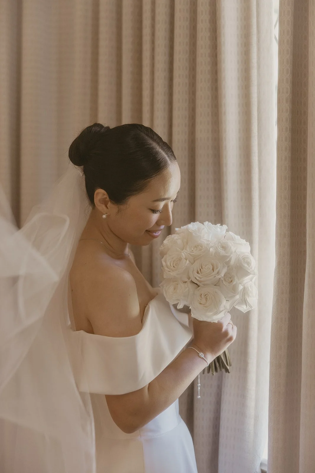 A bride in an off-the-shoulder wedding dress holding a bouquet of white roses, standing near a window with beige curtains, smiling softly.