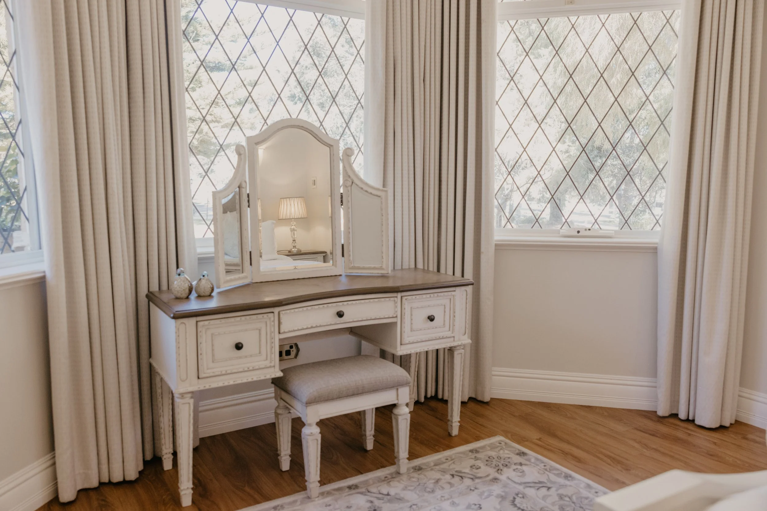 A white vintage vanity table with a matching stool in a room with large windows covered by striped curtains. The vanity has a mirror and a small table lamp.
