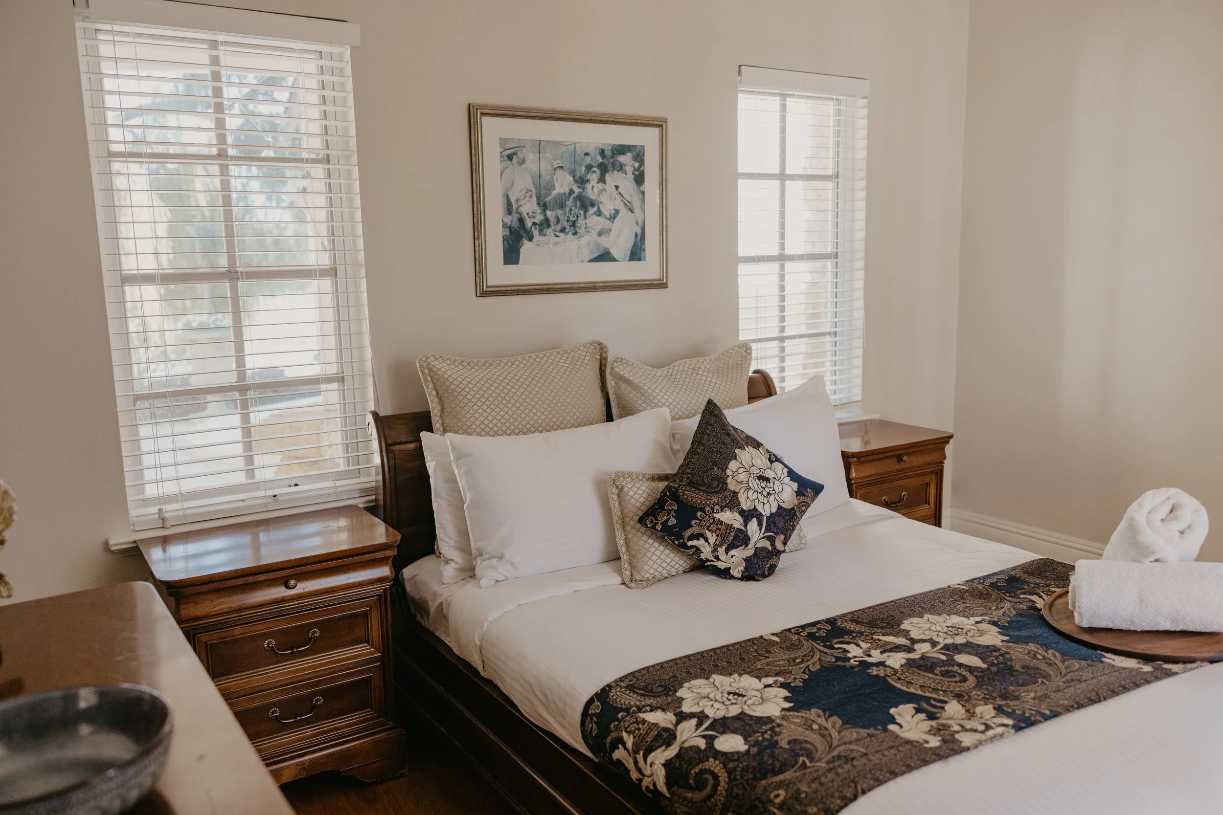 A neatly made bed in a bedroom with two windows, a framed artwork on the wall, and wooden bedside tables.