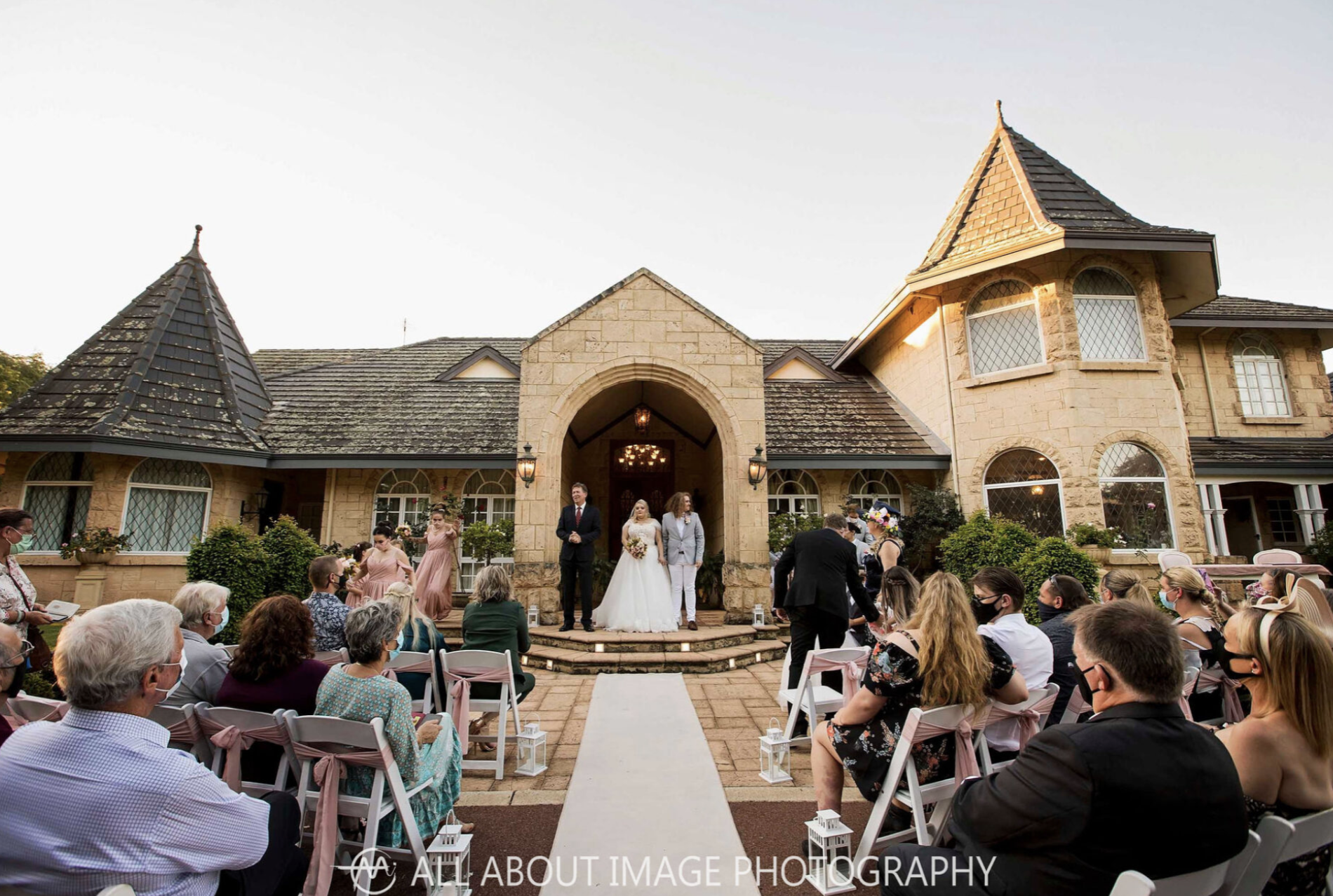Outdoor wedding ceremony in front of a large stone house with pointed towers. The bride and groom are standing on steps with guests seated on either side. The scene is during the evening with warm lighting.
