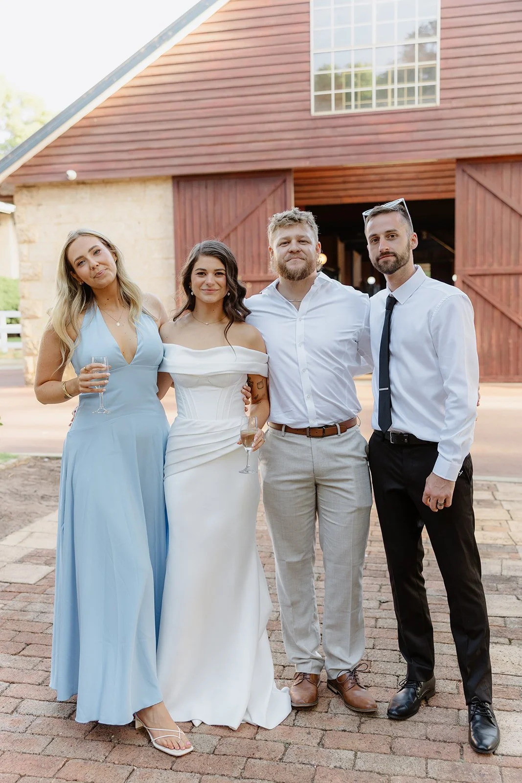 Four people standing outdoors in front of a barn, dressed in formal attire, holding wine glasses, and smiling at the camera.