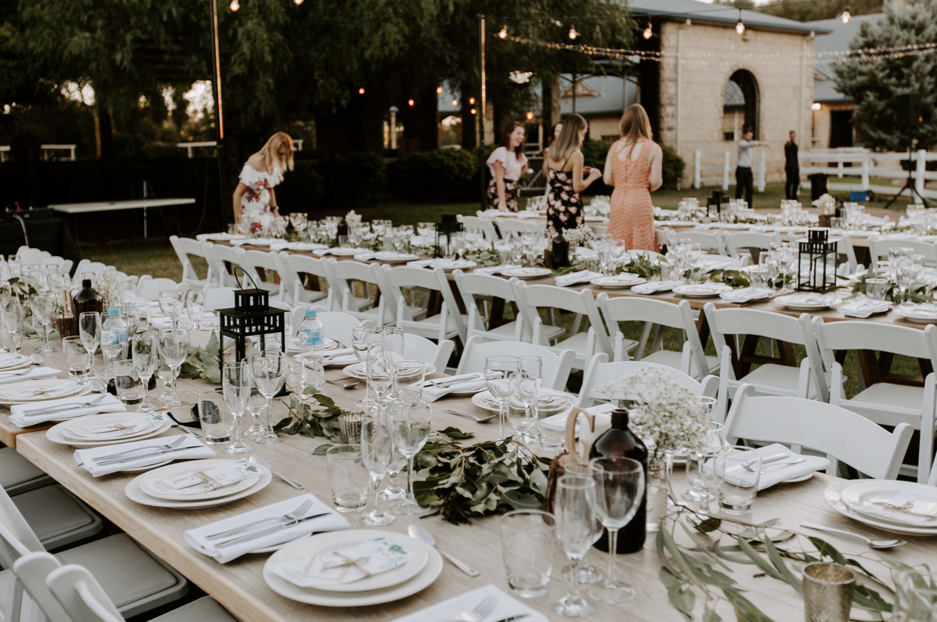 Outdoor wedding reception with long decorated tables, white chairs, and floral centerpieces at sunset.