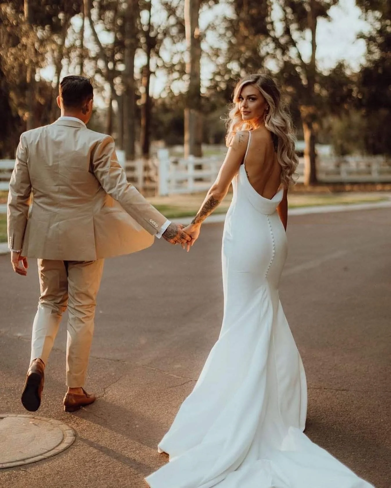 A bride in a long, white wedding dress holding hands with a groom in a light-colored suit as they walk outdoors during sunset.
