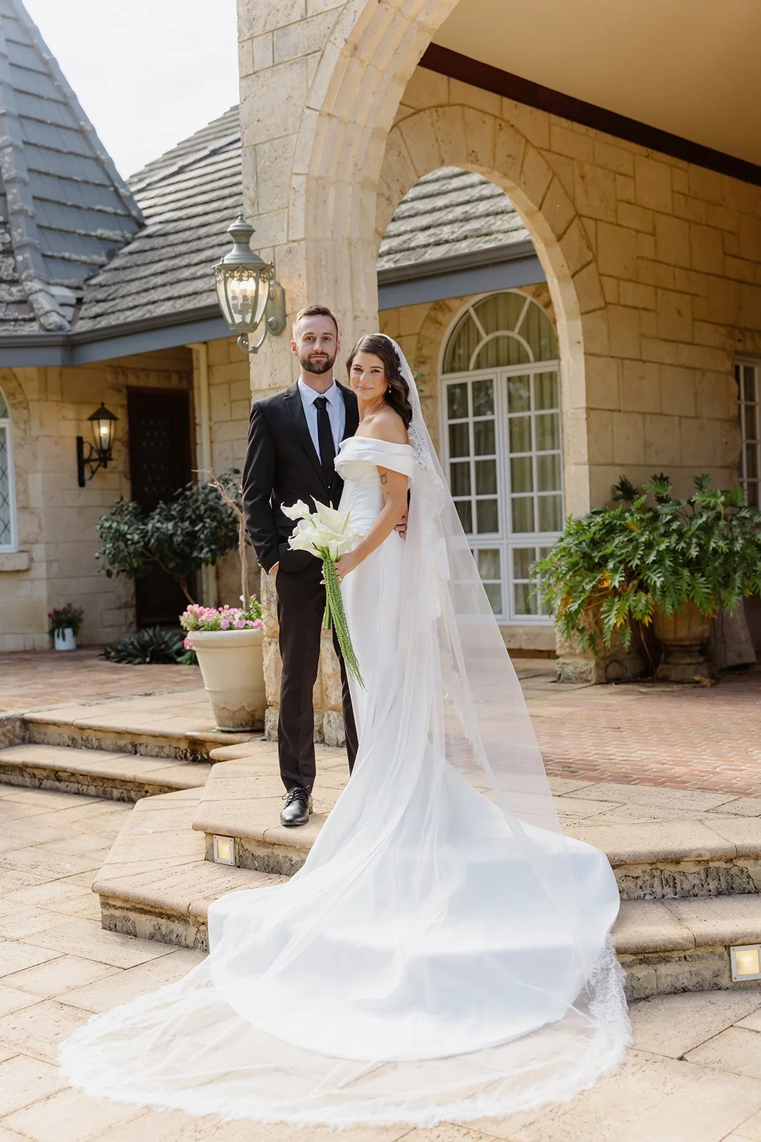 A newlywed couple standing on steps outside a stone building, dressed in wedding attire. The bride holds a bouquet of white calla lilies and wears a white off-the-shoulder wedding gown with a long train and veil. The groom wears a black suit, white s