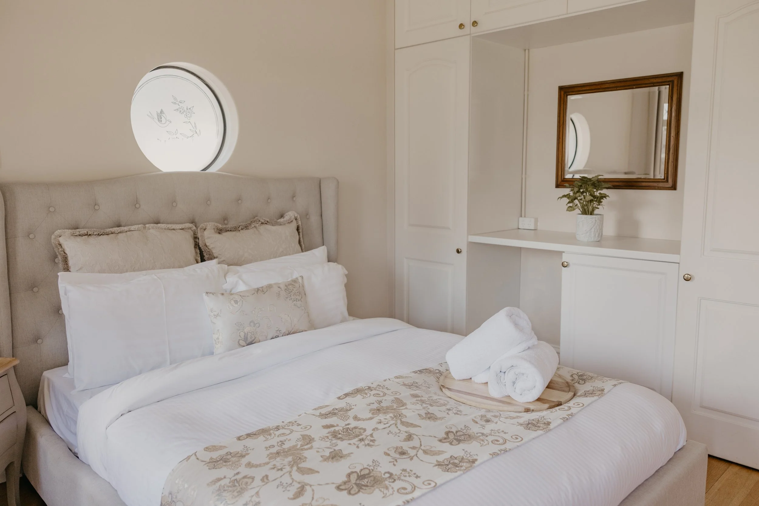 Made bed with white linens, beige decorative pillows, and a floral beige throw blanket. Rolled white towels on a wooden tray at the foot of the bed. Beige tufted headboard. White built-in cabinets and a mirror on the wall behind the bed. Small potted