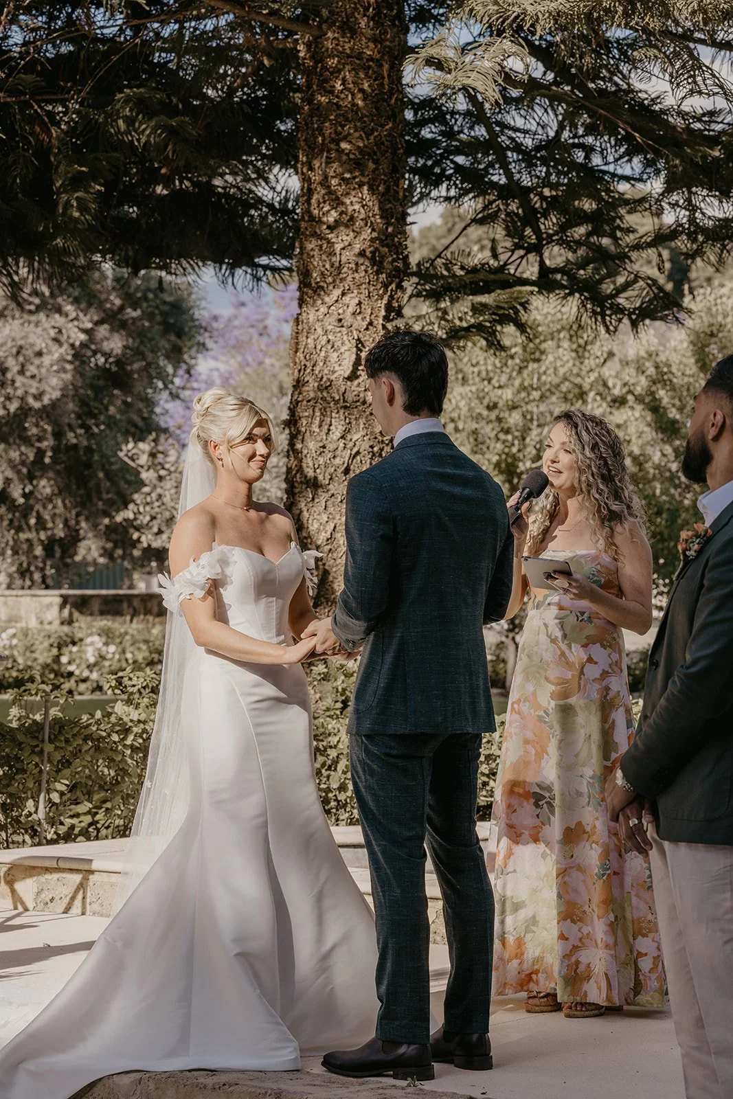 A wedding ceremony outdoors with a bride and groom holding hands, facing each other, under a large tree. The bride wears a white wedding dress with off-the-shoulder sleeves and a veil. The groom wears a dark suit. An officiant with curly hair holds a
