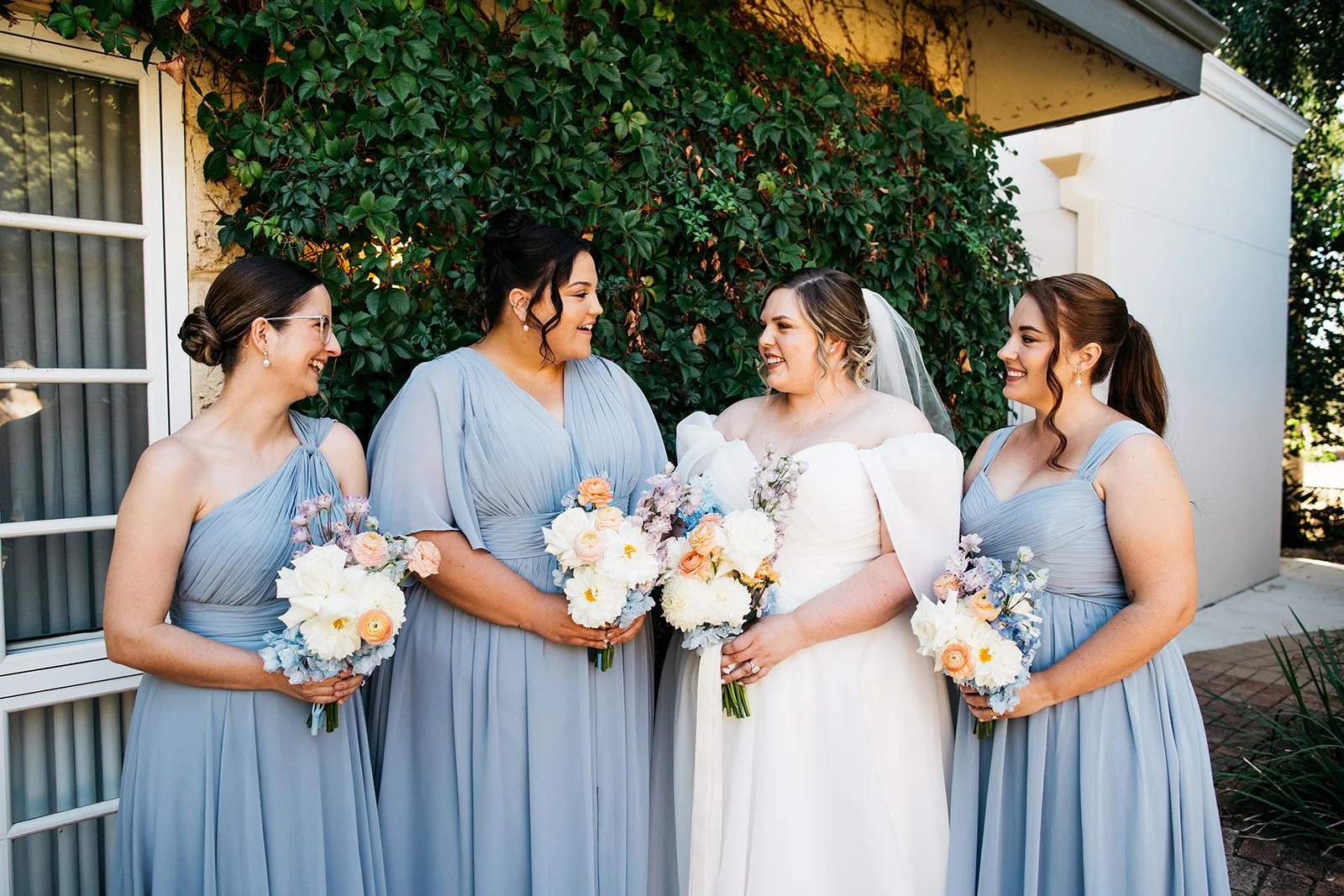 A bride and her four bridesmaids standing outside, smiling and holding bouquets of flowers, with greenery and a building in the background.