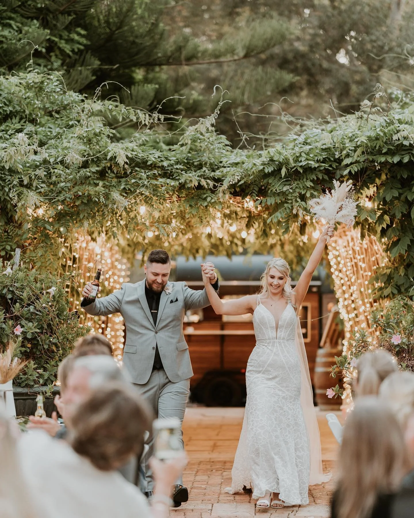 A newlywed couple dancing at their wedding reception under a leafy archway decorated with string lights. The groom is wearing a light grey suit and the bride is holding a bouquet with one arm raised.