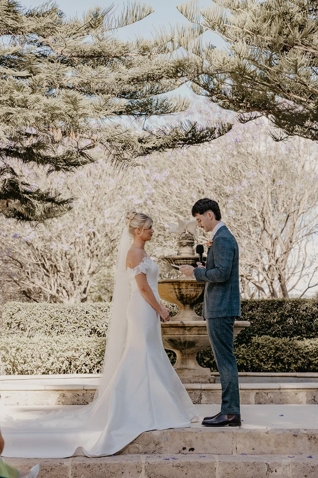 A wedding ceremony outdoors with a bride and groom standing in front of a fountain, surrounded by trees and bushes, during daytime.