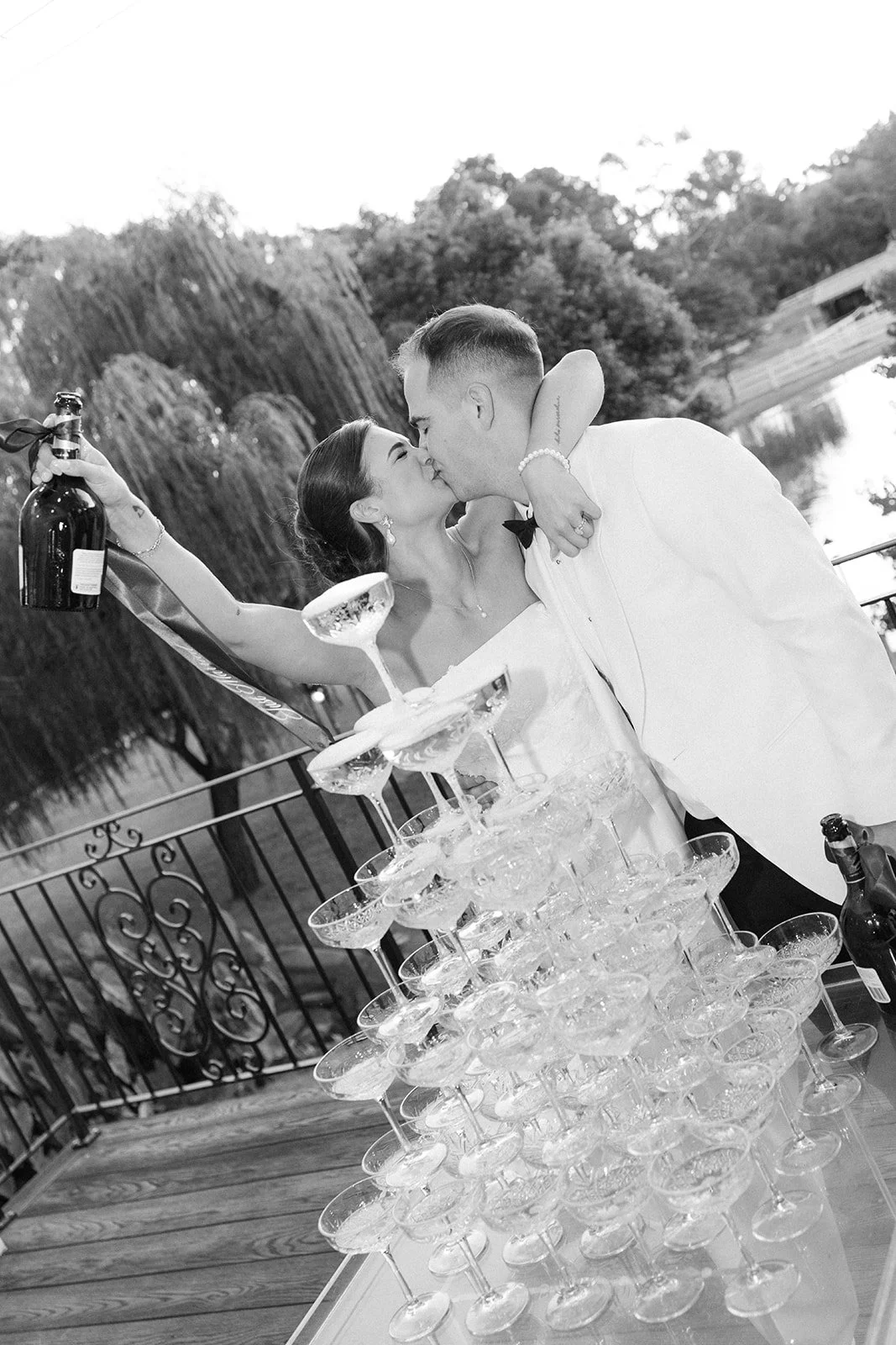 A black and white photo of a newlywed couple sharing a kiss at their wedding reception. The bride is holding a bottle of champagne and has her arm around the groom. There is a tower of empty champagne glasses in front of them on a table, and the back