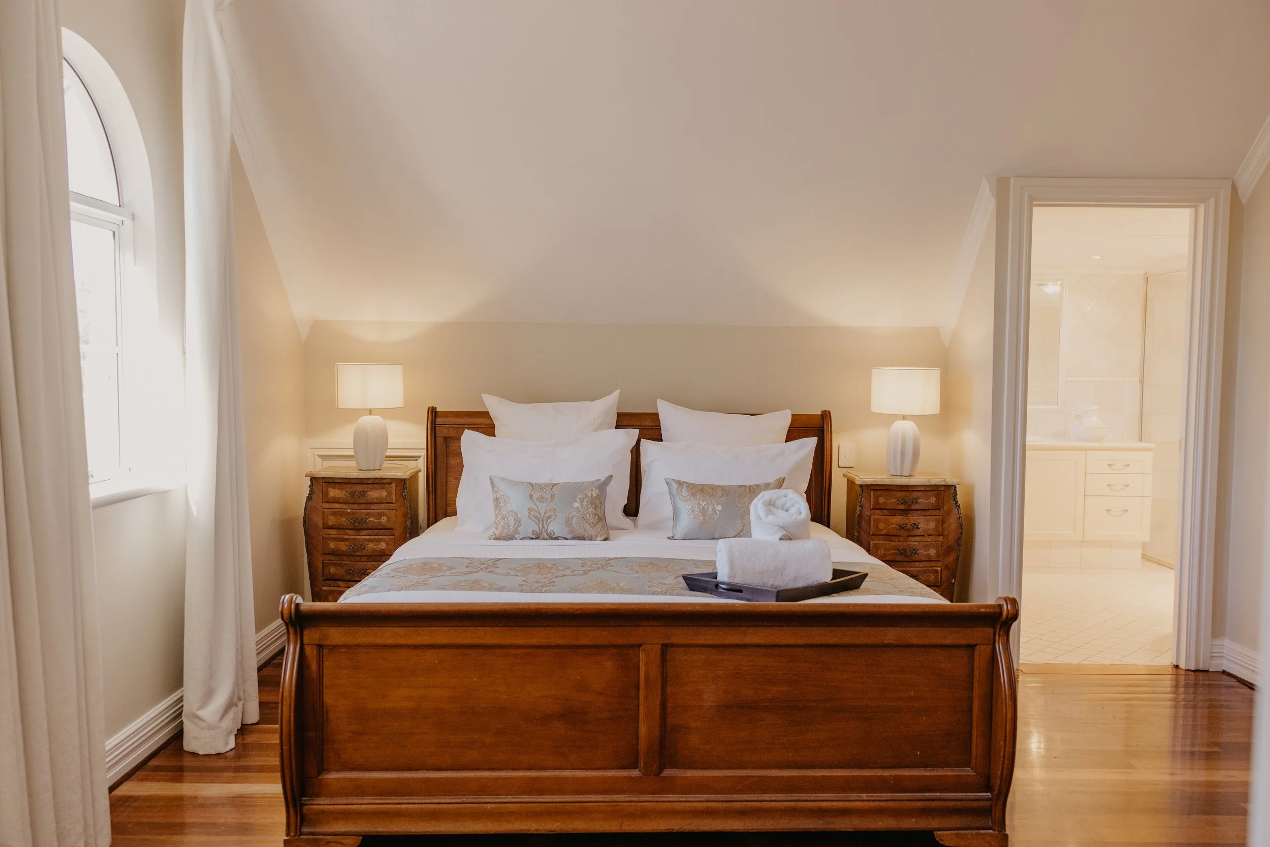 A cozy bedroom with a wooden bed frame, neatly made with white and beige bedding, surrounded by two nightstands with white lamps, and natural light coming through a window with white curtains. There is an open door leading to a bathroom.