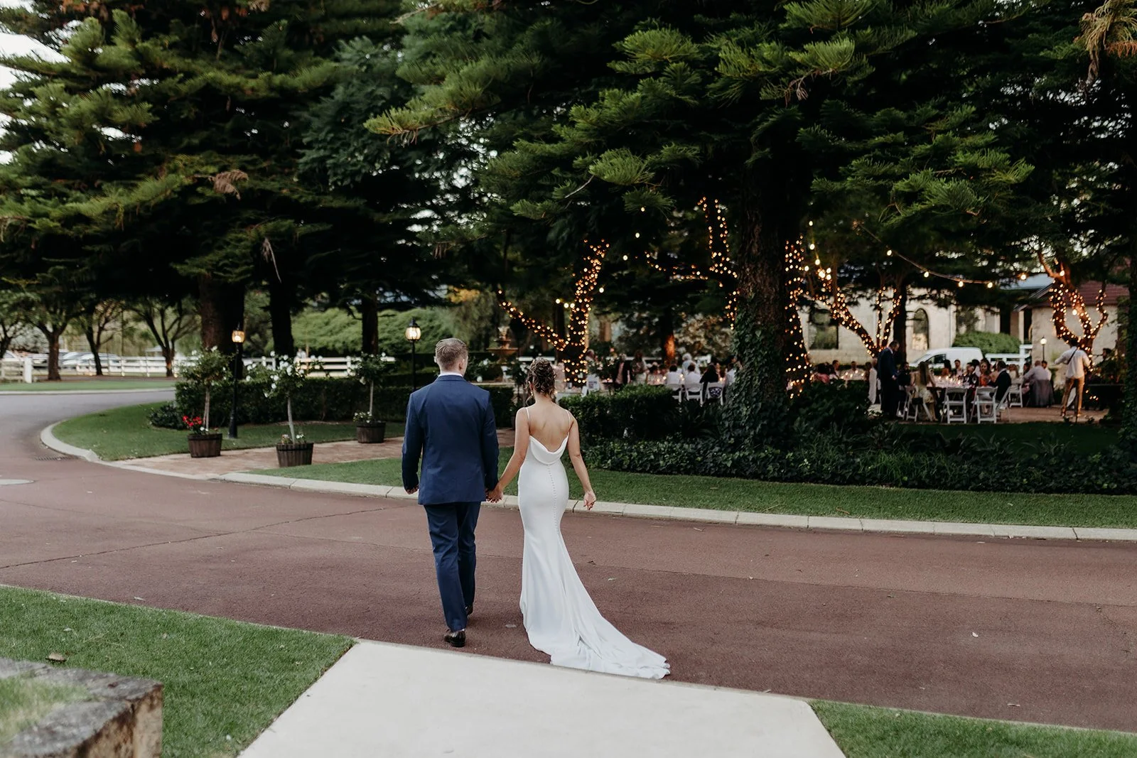 Couple holding hands walking towards outdoor wedding reception under trees decorated with string lights, at dusk.
