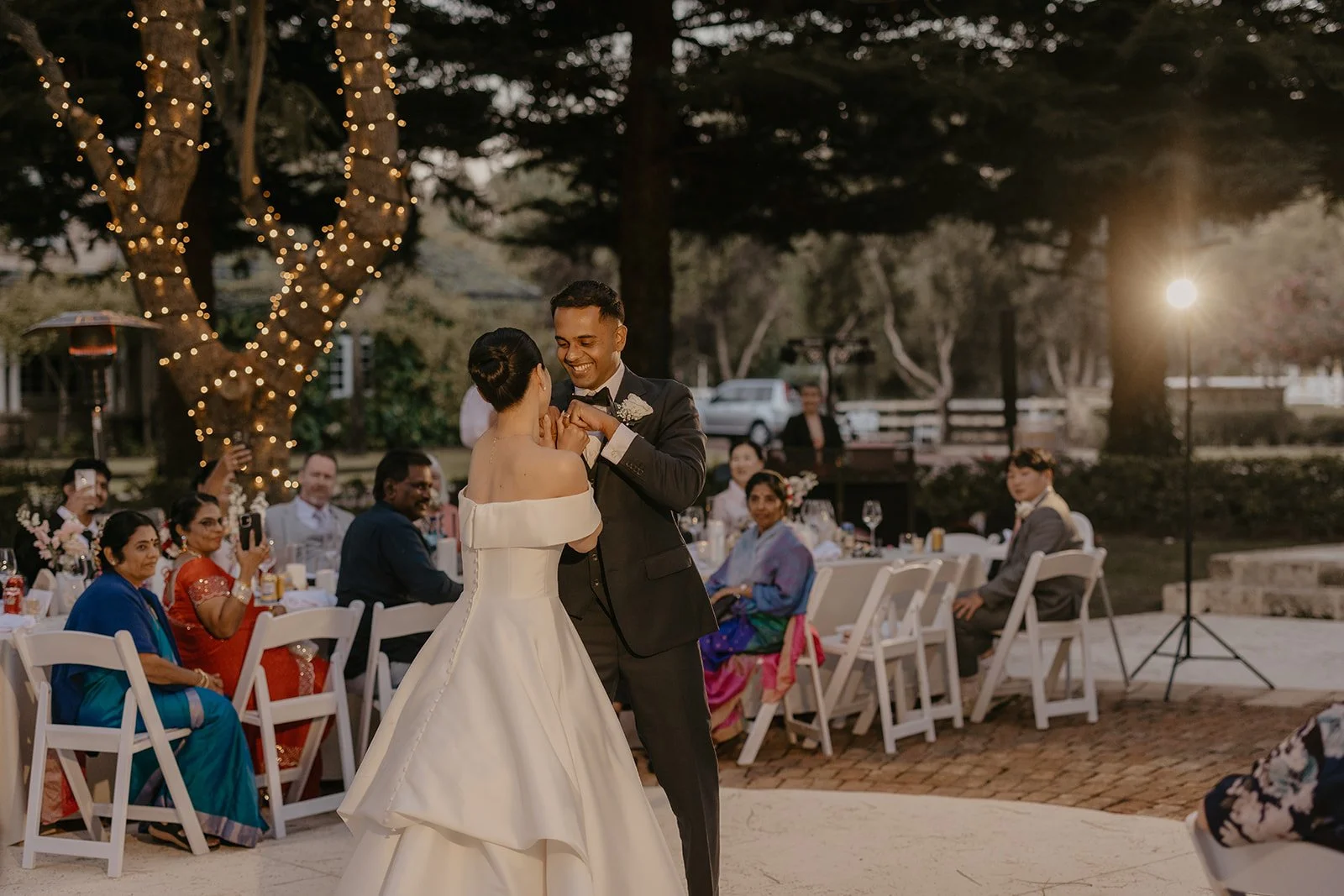 A bride and groom dance at their wedding reception outdoors during sunset. The bride wears a white off-the-shoulder gown with a long train, and the groom wears a black tuxedo. Guests sit at decorated tables, some watching and taking photos. String li