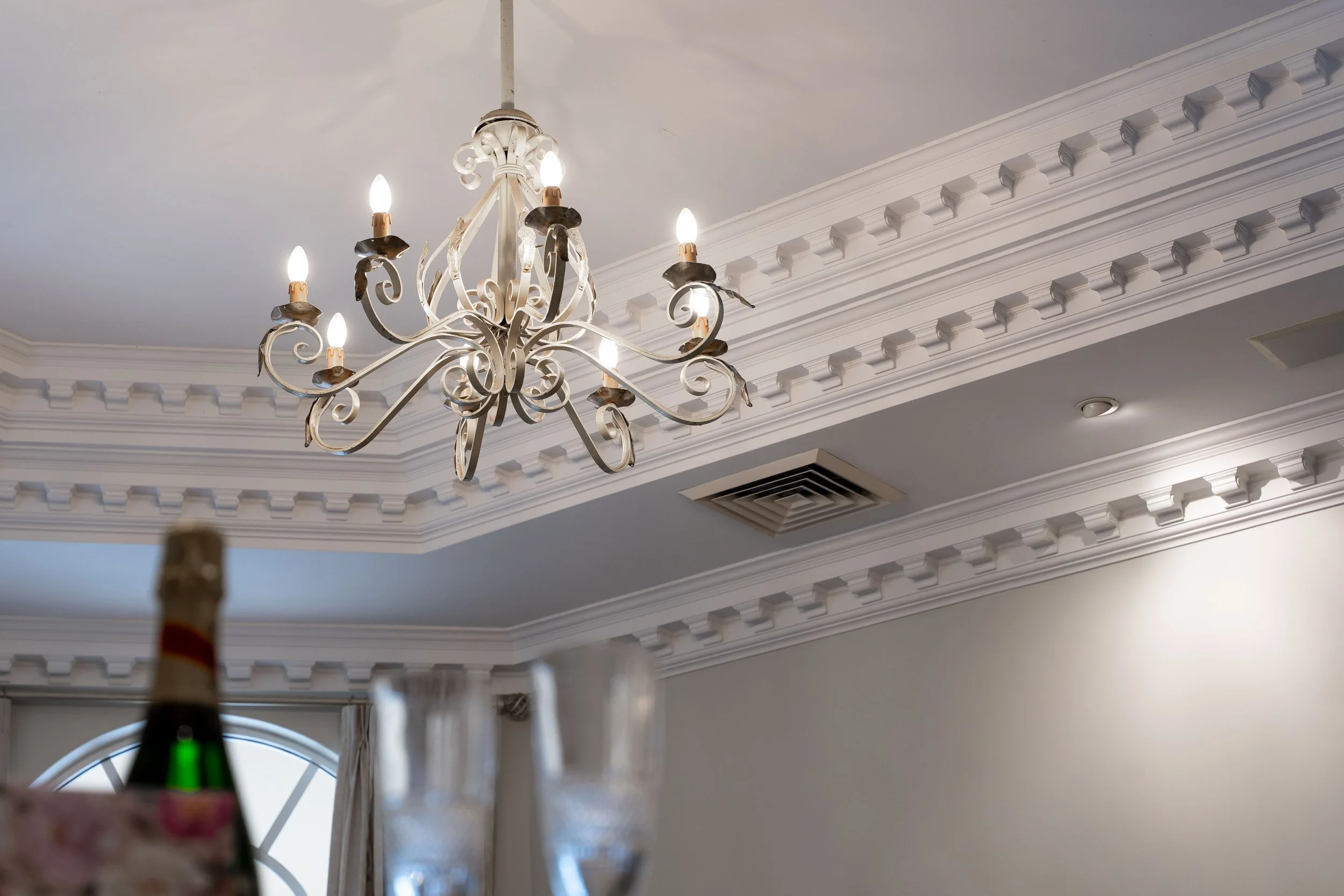 A chandelier hanging from the ceiling with decorative crown molding. Part of a wine bottle is visible in the foreground, and a large circular window with curtains is in the background.