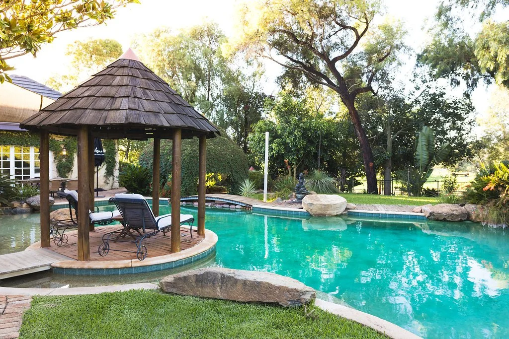 A backyard swimming pool with a small wooden gazebo and lounge chairs, surrounded by trees and lush greenery.