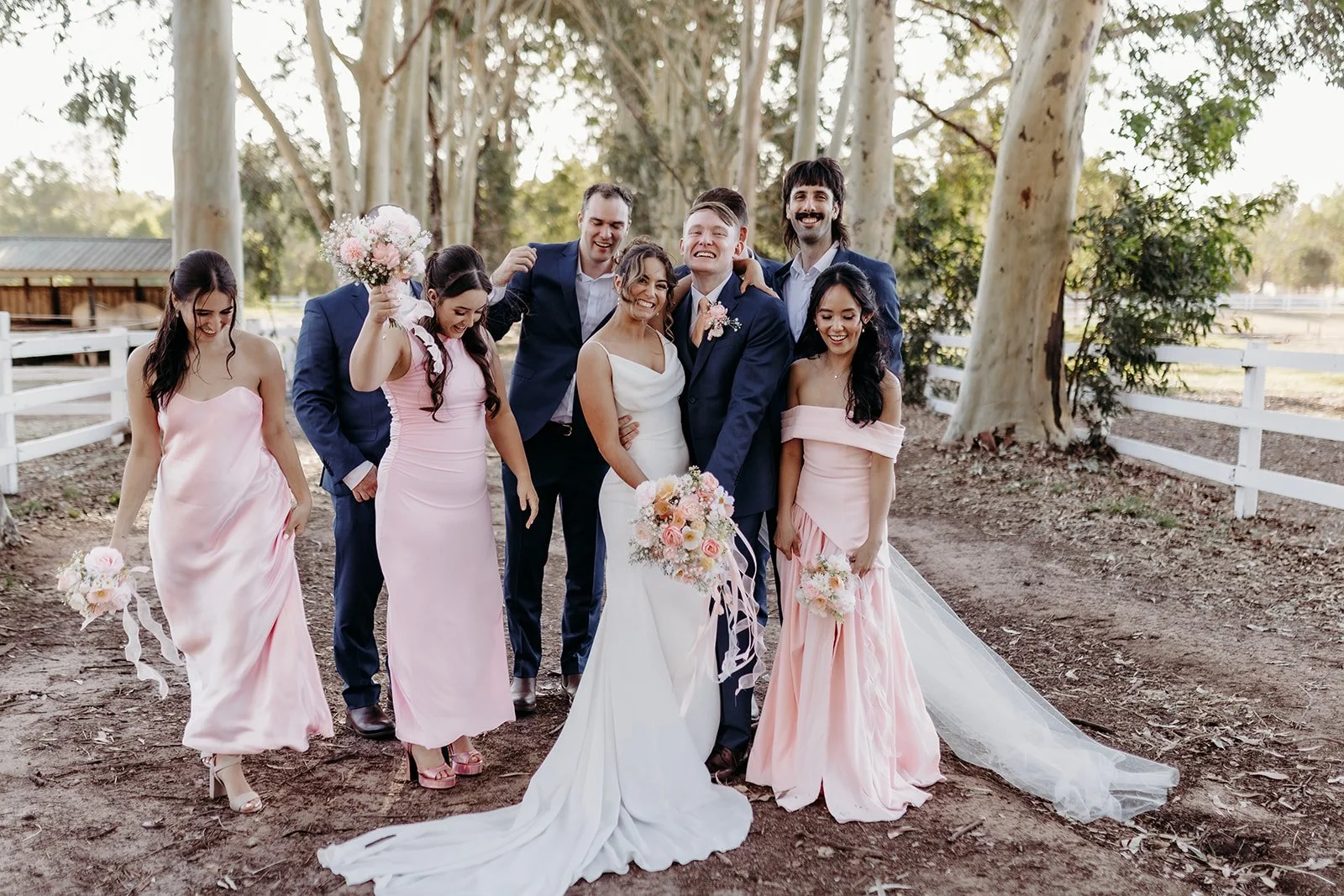 A wedding party of eight people standing outdoors among tall trees, with the bride and groom at the center, all smiling and dressed in wedding attire; the bride in a white gown and the groom in a navy suit, with bridesmaids in pink dresses and grooms