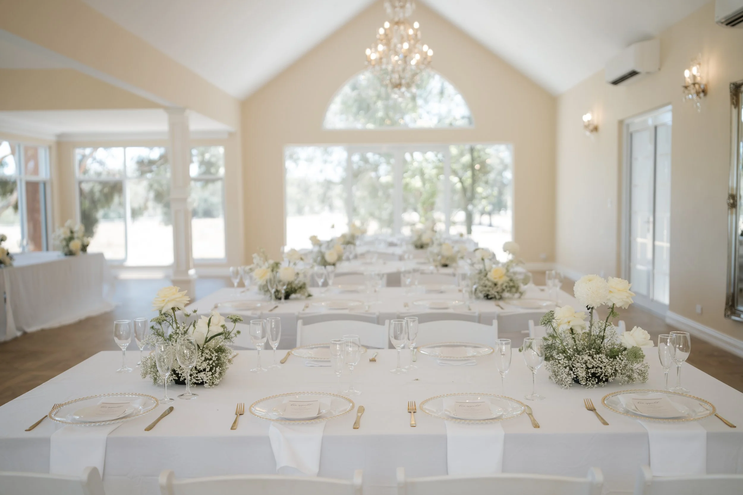Elegant banquet hall with high v-shaped ceiling, large arched window, chandelier, wall sconces, and air conditioning units, prepared for a wedding or formal event.