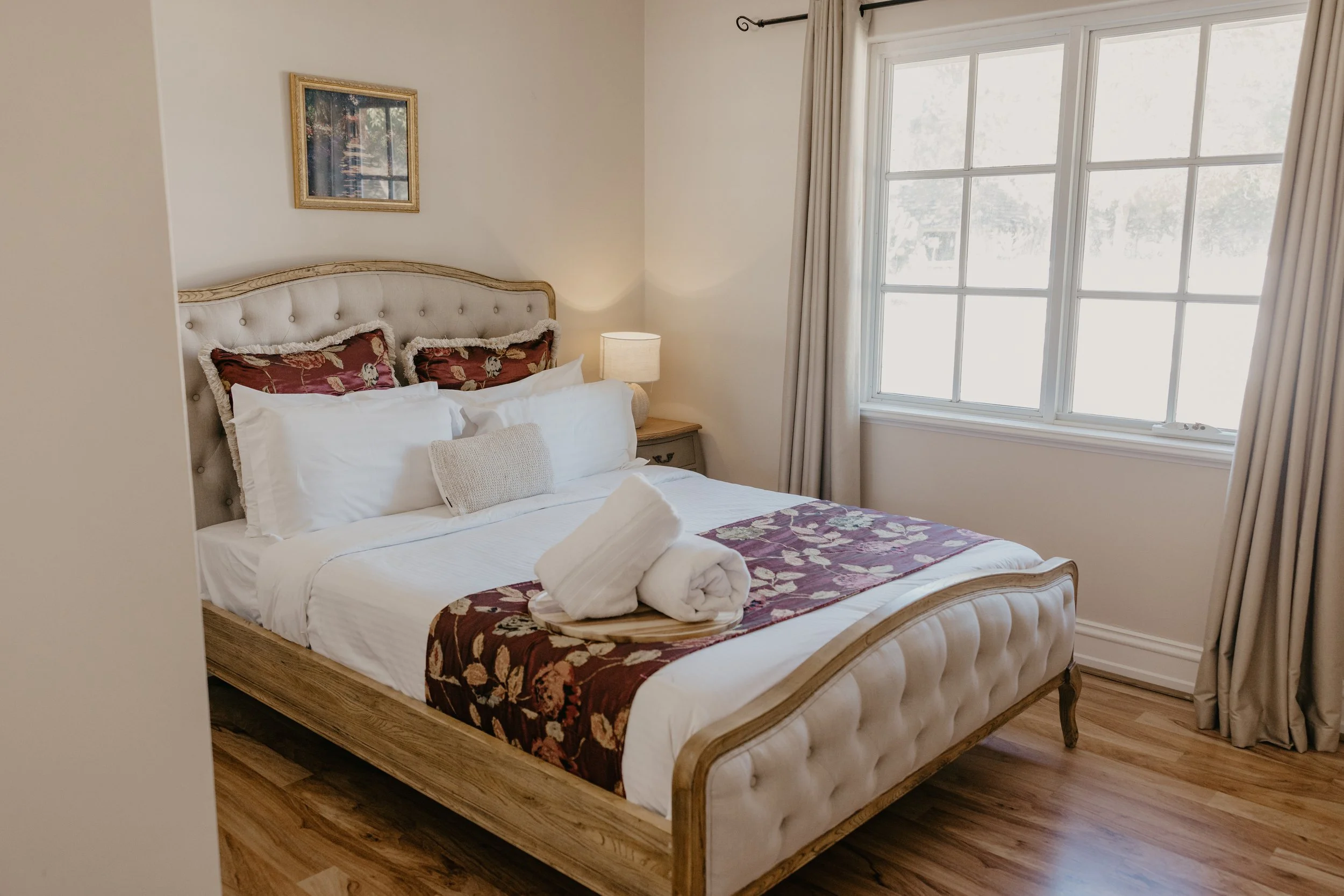 A cozy bedroom with a wooden bed frame, white bedding, and decorative pillows with floral patterns. There is a bedside table with a lamp and a rolled towel on the bed. A large window with white curtains lets in natural light, and a framed picture han
