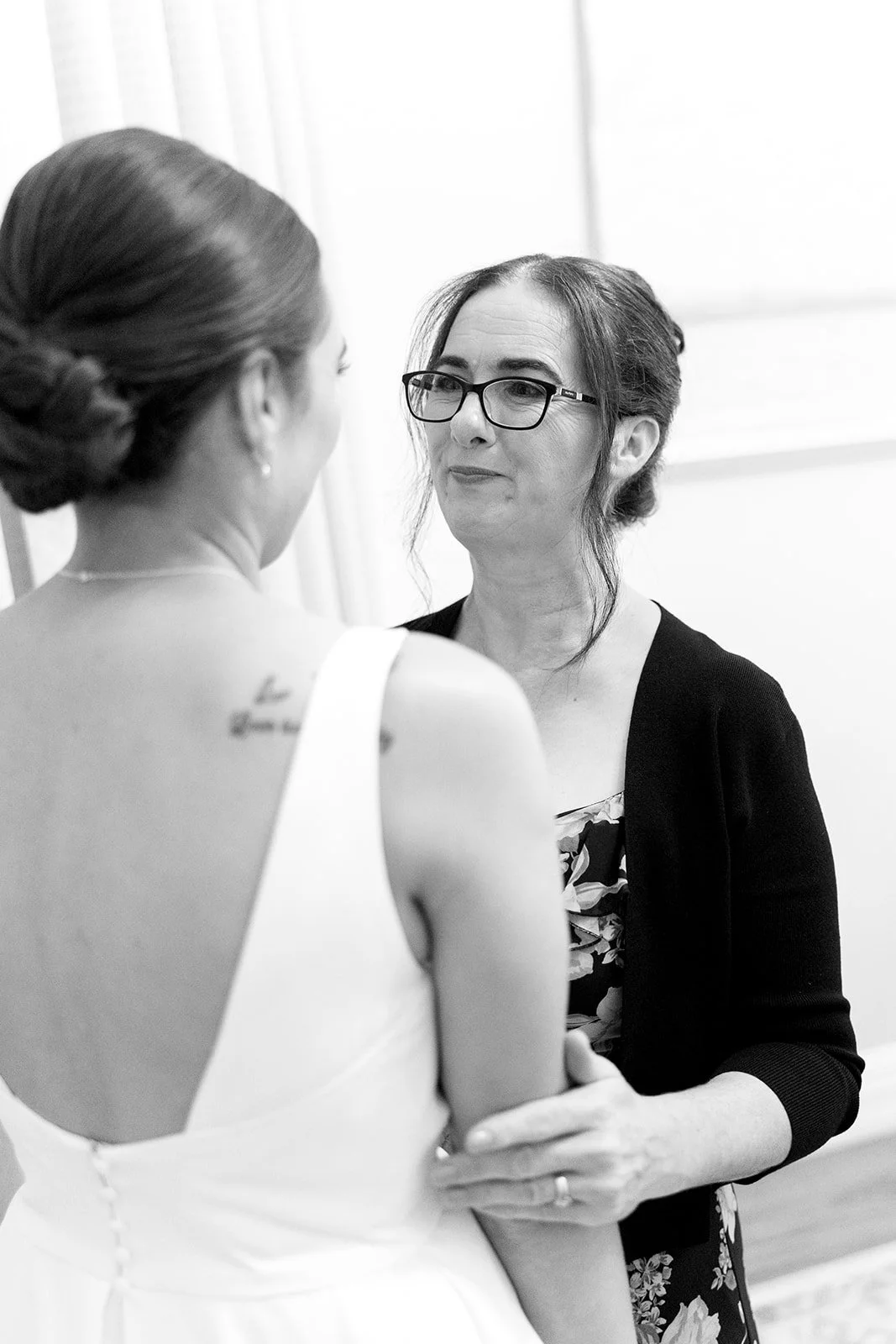 A woman in a white dress with her hair up, facing another woman with glasses, dark hair, and a black cardigan, who is holding her hand.