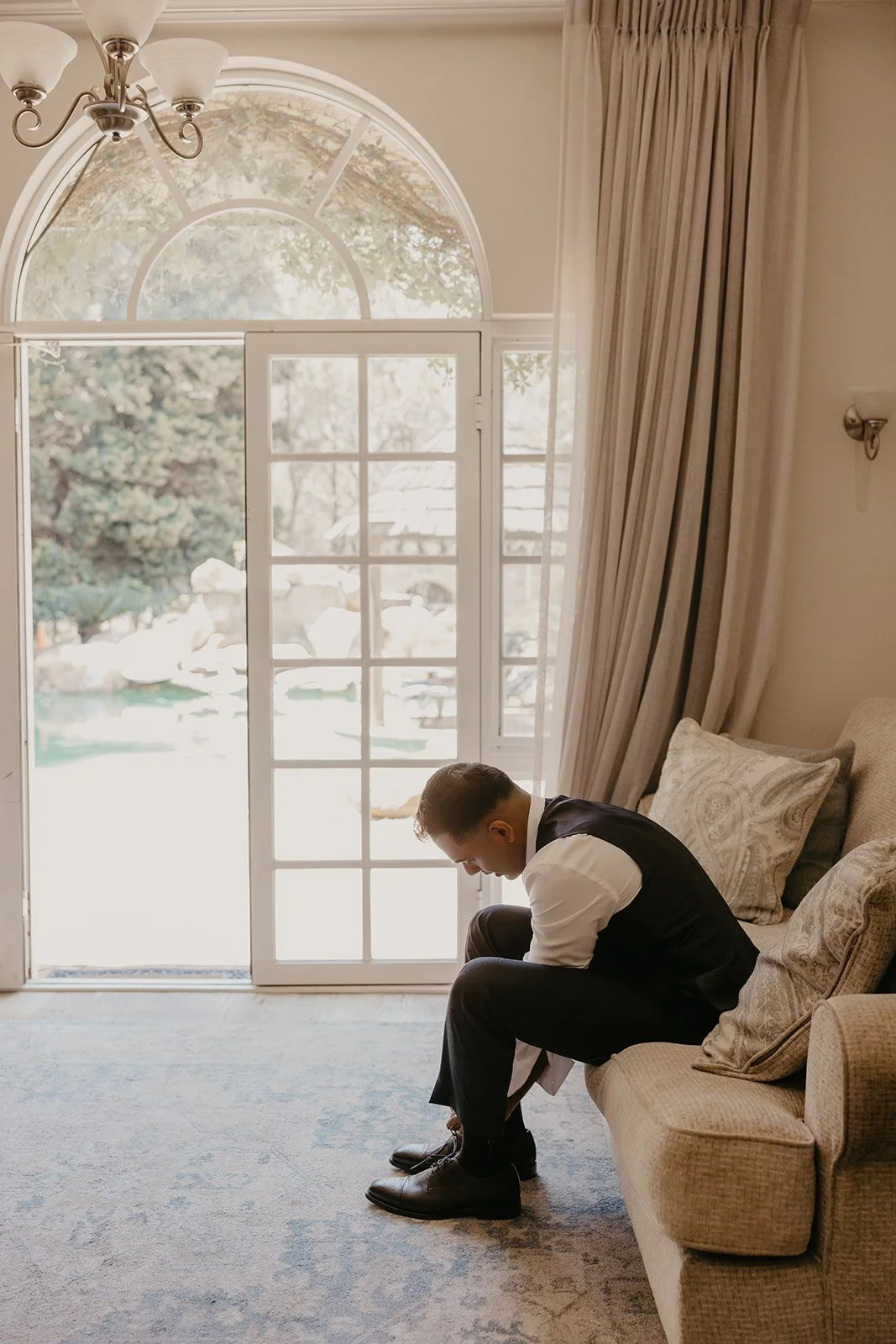 A man in formal attire, sitting on a beige sofa, bending forward to tie his shoe, with a large window and curtains behind him.