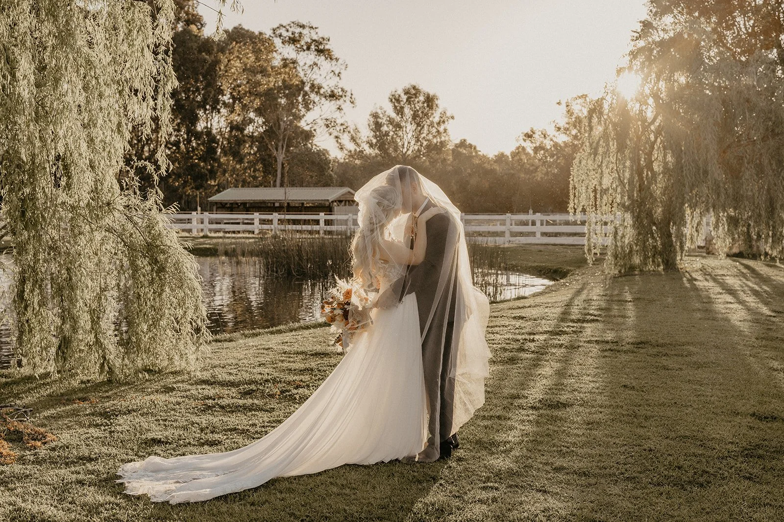 A bride and groom embrace in a sunset outdoor setting, with trees and a pond in the background in the Swan Valley Perth at Brookleigh Estate