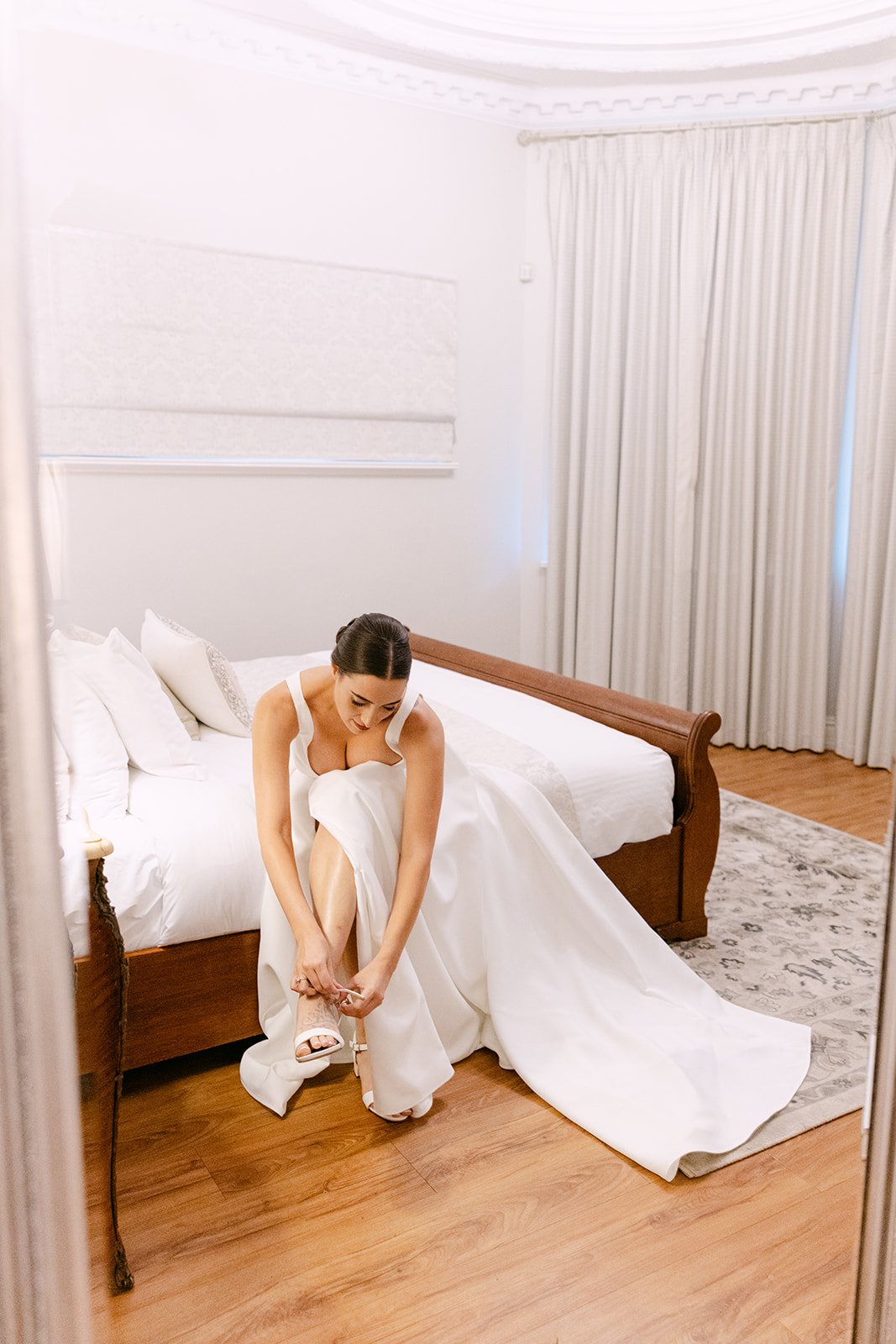 A woman in a white wedding dress sitting on a bed and putting on high-heeled shoes in a bedroom with white curtains, a wooden bed frame, and a patterned carpet.