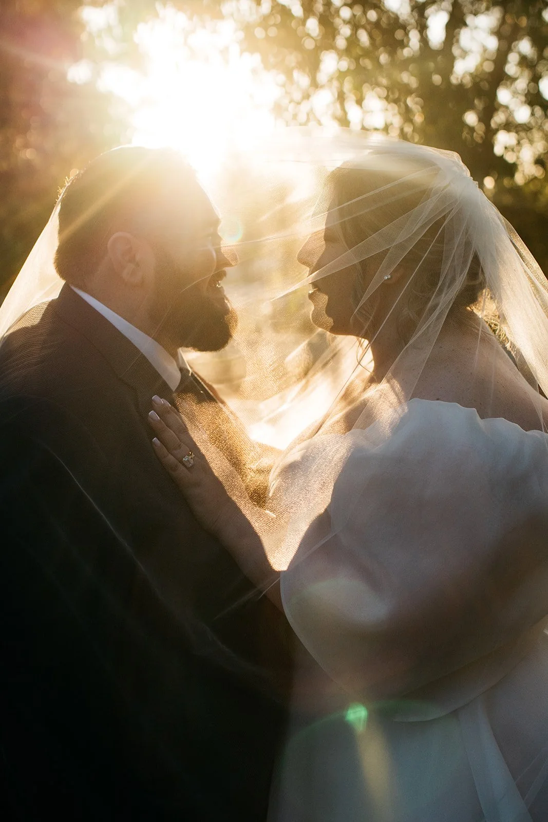 A bride and groom facing each other under a wedding veil, with sunlight creating a backlit glow and lens flare, outdoors surrounded by trees.