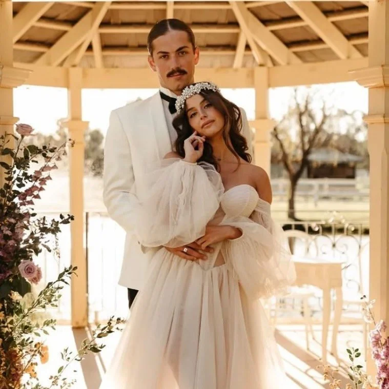 A man and woman in wedding attire standing together under a wooden gazebo. The man is in a white tuxedo and the woman is in a strapless wedding gown with a flower crown. They are outdoors with trees and a body of water in the background.
